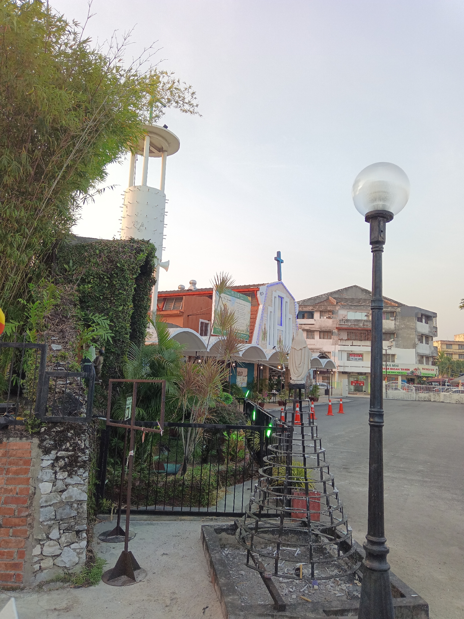 The image captures the exterior of a church located in Klang City, Malaysia. The church features a white facade adorned with a blue cross and decorative lights. In front of the church, a statue of a person, possibly a religious figure, stands near orange traffic cones. A tall lamppost with a spherical light fixture is prominent in the foreground. To the left of the church, a white tower structure rises, surrounded by lush green bamboo plants and ivy-covered walls. A black metal fence encloses a garden area with various plants and a potted plant. In the background, several multi-story buildings are visible, one of which displays signage in what appears to be Malay. The overall setting suggests a public space within the city.