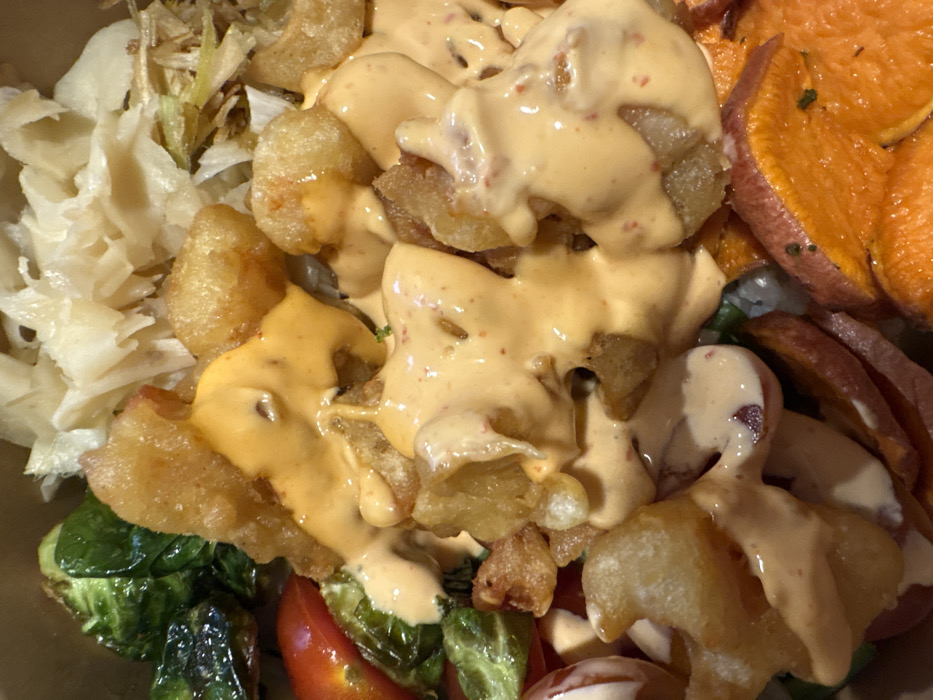 A close-up shot features a food bowl filled with a variety of ingredients. There are fried, bite-sized pieces coated in a creamy, orange-tinted sauce with reddish specks. Accompanying these are sliced sweet potatoes, roasted brussel sprouts, shredded cabbage, and red cherry tomatoes, offering a colorful and diverse culinary experience.
