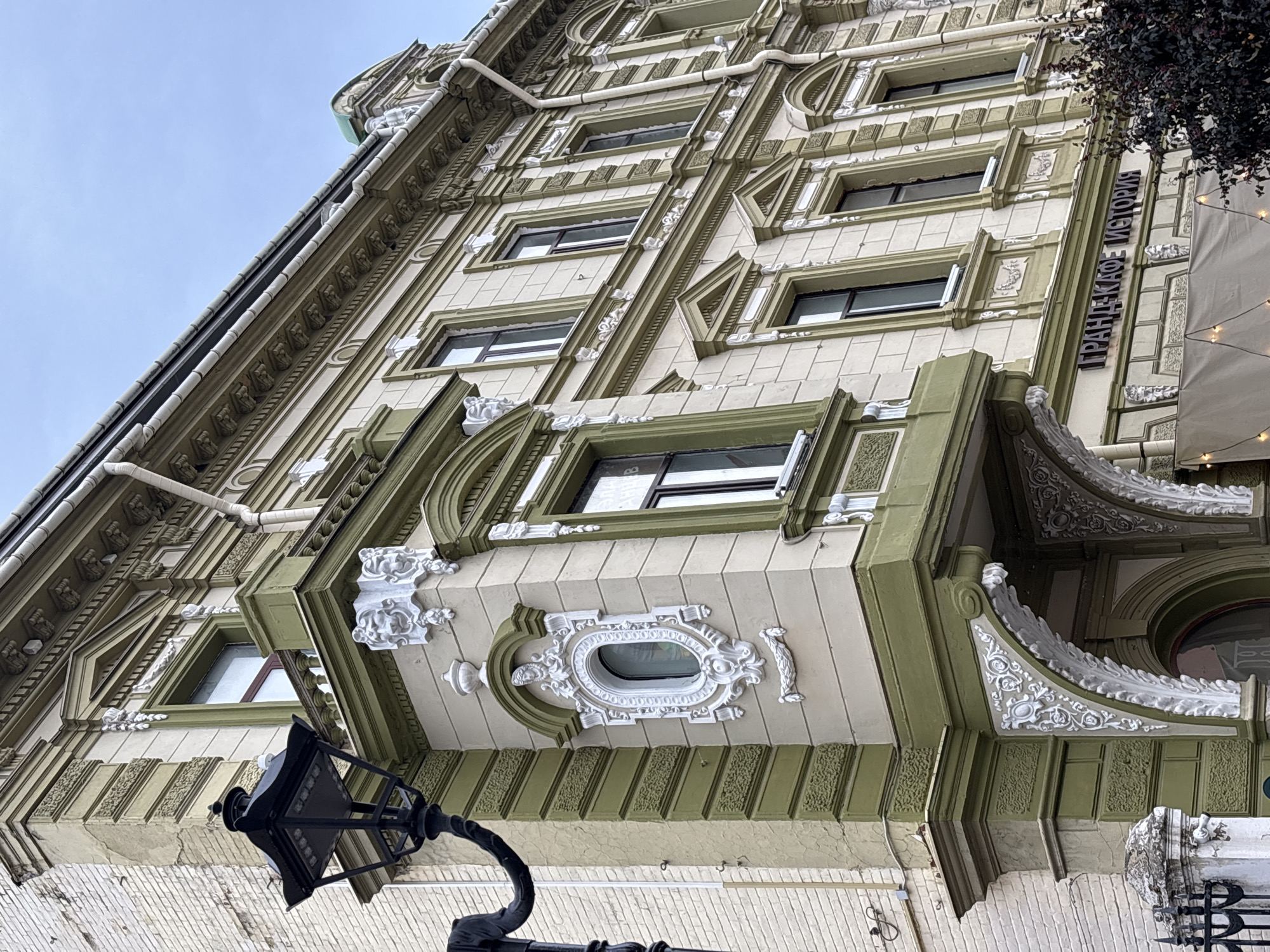 The image shows a detailed view of the facade of a historic building in Nizhny Novgorod, Russia. The building is adorned with intricate architectural details, including ornate window frames, decorative moldings, and a bay window. The dominant colors are a light cream and a muted green. A black lamppost is visible on the left side of the image, and a portion of a dark-leaved tree is on the right. The sky above is overcast. The text "ГРАНДЪ КАФЕ ИСТОРІЯ" (Grand Cafe Istoriya) is visible on the building, indicating the name of the establishment. The architectural style suggests it is from the late 19th or early 20th century.