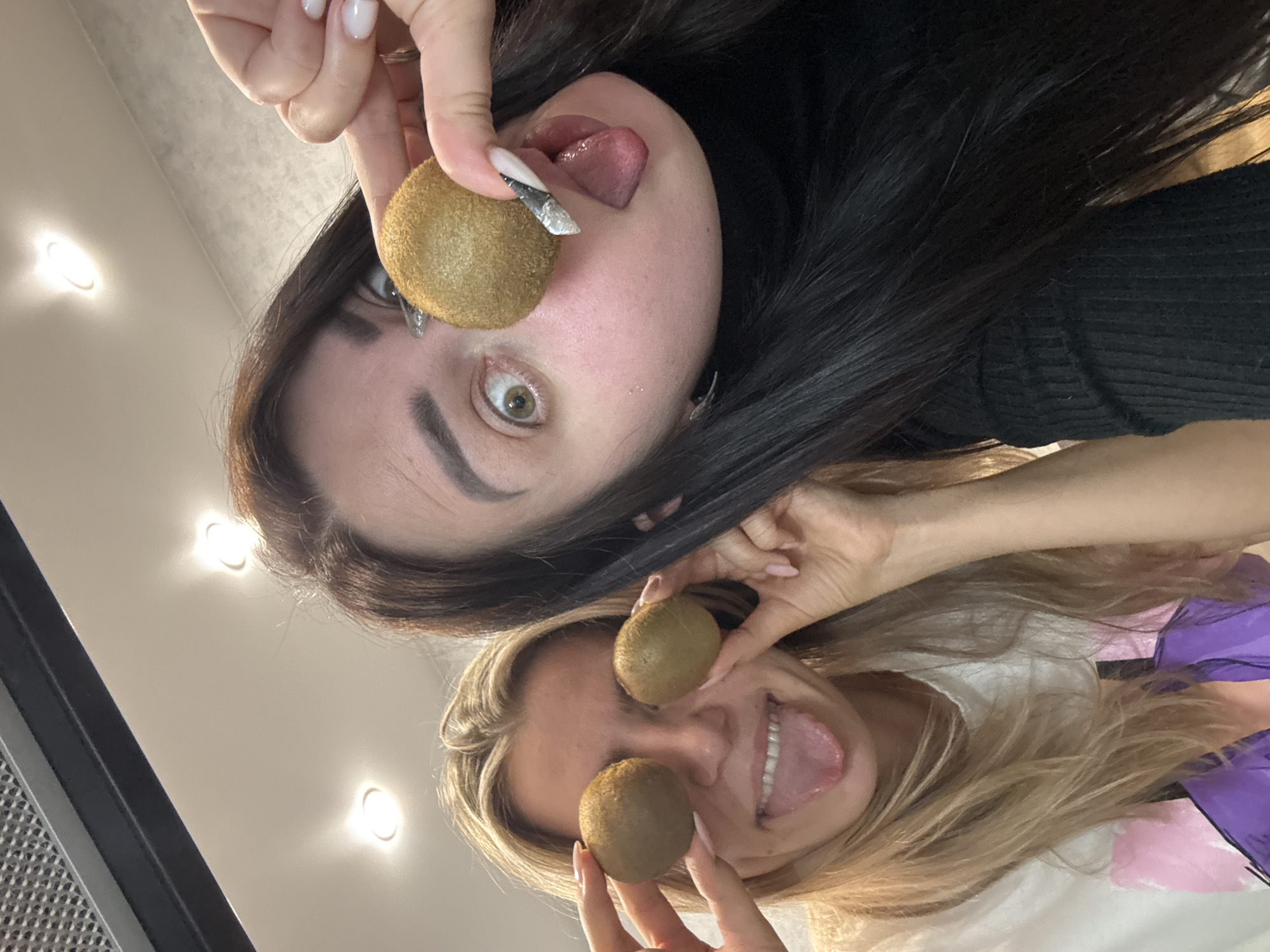 An overhead shot captures two young women playfully posing with kiwis, viewed from a low angle looking upwards, making them appear inverted. The woman at the top of the frame has long dark hair, prominent dark eyebrows, and striking light-colored eyes, one of which is visible and wide open. She wears a dark, possibly black, ribbed long-sleeved top. With manicured nails, one adorned with a glittery design, she holds a kiwi near her eye while her tongue is playfully extended from her mouth.
Below her, another woman with long blonde hair smiles widely, her tongue also extended. She holds two kiwis, one over each eye, resembling oversized spectacles. She appears to be wearing a white top with hints of a purple and pink pattern visible around her shoulders. Her nails are also manicured, painted a light pink.
The background consists of a plain light-colored ceiling, illuminated by multiple bright, recessed spotlights. On the far left, a section of what appears to be a black range hood or ventilation grate is visible. The overall mood is lighthearted and humorous. There is no visible text in the image.