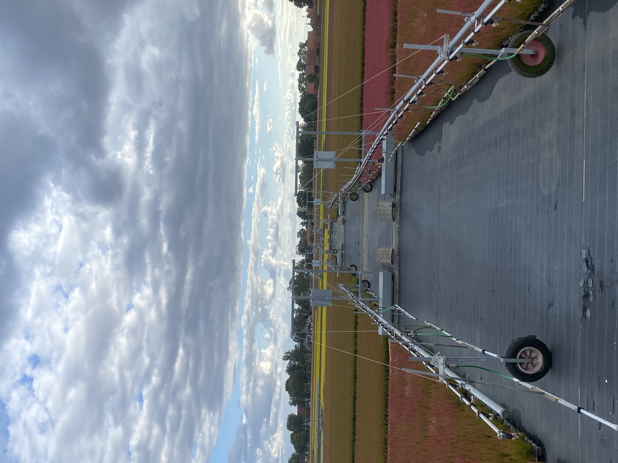 This is a high-angle, wide shot of a nursery or agricultural field in Schwarme, Germany. The scene is dominated by rows of young plants, colored red and green, stretching into the distance. Overhead, a pathway is defined by two parallel, wheeled irrigation or spraying systems. These systems, made of metal poles and pipes, are positioned over the dark, textured ground cover. The sky above is filled with dramatic clouds, with patches of blue sky visible. In the far background, trees and buildings can be seen, suggesting the edge of a town or rural development. The overall impression is one of a managed, cultivated landscape under a dynamic sky.