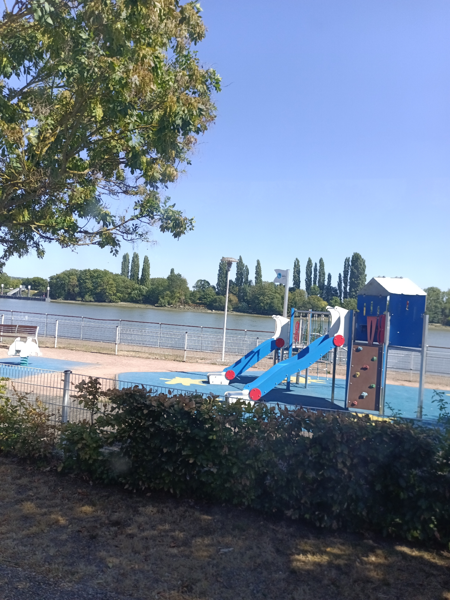 This image depicts a children's playground situated along the banks of the Seine River in Duclair, France. The playground features a blue and red multi-level play structure with slides and a climbing wall. A blue rubber safety surface is visible beneath the equipment, interspersed with yellow star-shaped designs. In the background, the tranquil Seine River flows, bordered by a tree-lined embankment on the opposite shore. A white metal railing separates the playground from a paved walkway and a grassy area in the foreground. The sky is a clear, bright blue.