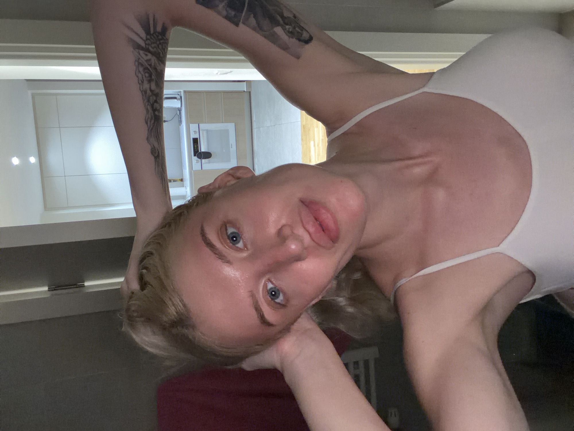 A fair-skinned woman with blonde hair and blue eyes is in the frame, wearing a light-colored tank top. She has tattoos on her upper arms. The background shows a portion of a kitchen, identifiable by white cabinets and a microwave. Given the metadata containing the city of Belgrade, Serbia, it can be inferred that this photo was taken indoors, presumably at the subject's residence in Belgrade. The woman is positioned at an unusual angle, tilting her head to the side. Her pose involves her arms being raised. The lighting is somewhat dim.