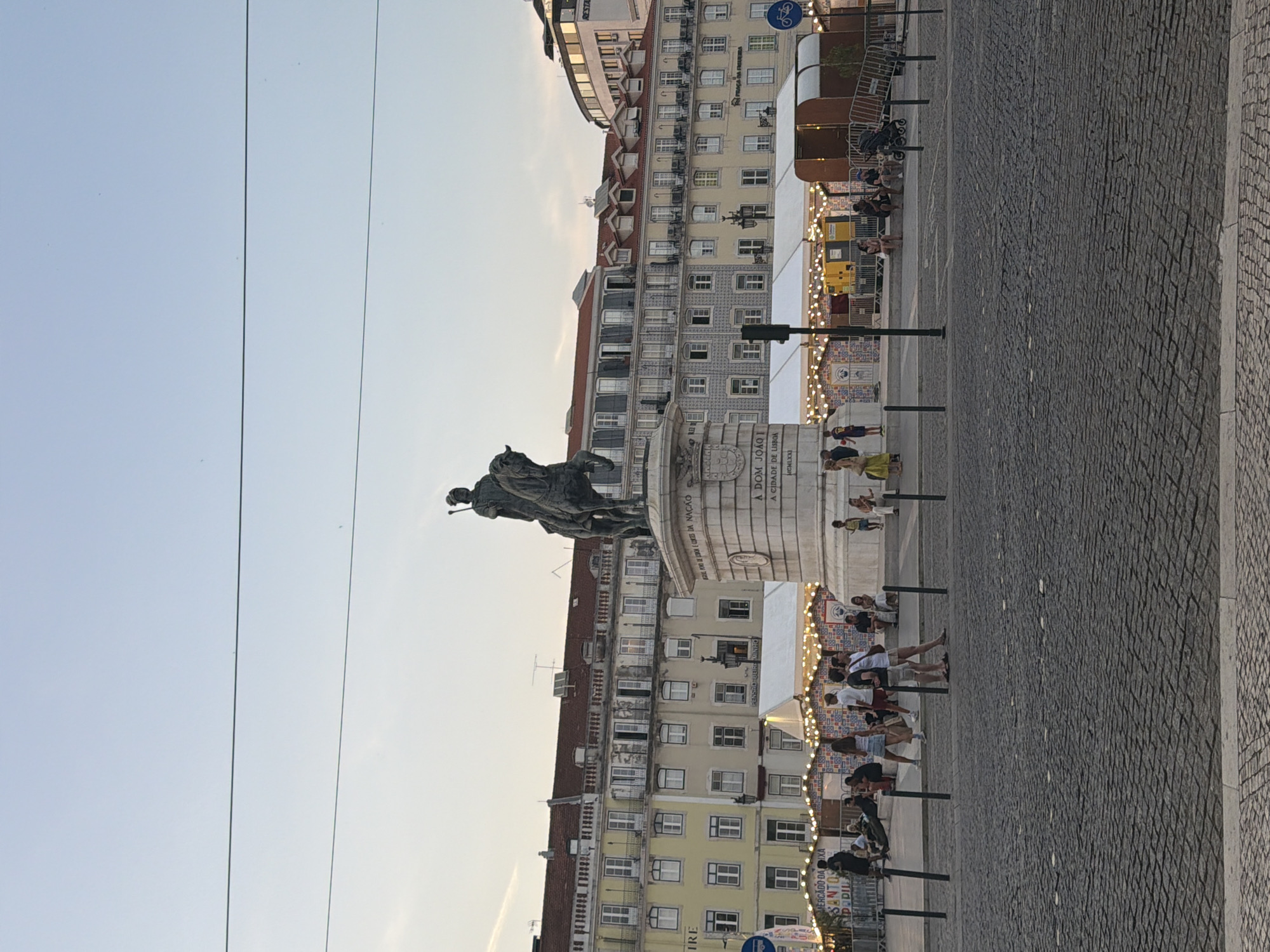 The image captures a wide shot of Praça do Comércio in Lisbon, Portugal, during what appears to be late afternoon or early evening. In the foreground, a cobblestone pavement fills the lower half of the frame. A large equestrian statue, likely of King José I, dominates the center of the image, mounted on a stone pedestal.
Behind the statue, a row of historic buildings with red tiled roofs and numerous windows stretches across the background. Some of these buildings are painted yellow, while others are a lighter beige or white. The architecture suggests classical European design.
In front of the buildings, several white tents with decorative string lights are set up, indicating a temporary market or event. Various people are visible, some standing and conversing, others sitting in small groups, and a few walking. The people are dressed in casual summer clothing. A stroller is visible near some seated individuals.
The sky above is a pale blue with a hint of orange, suggesting the time of day. Two thin dark lines, likely power lines or cables, traverse the upper portion of the image horizontally. On the right side, a modern-looking structure with a curved roof and signage that appears to read "RESTAUR..." is partially visible, contrasting with the older buildings. A blue traffic sign with a bicycle symbol is also visible on the right edge. The overall atmosphere is one of a public square bustling with activity during a pleasant time of day.