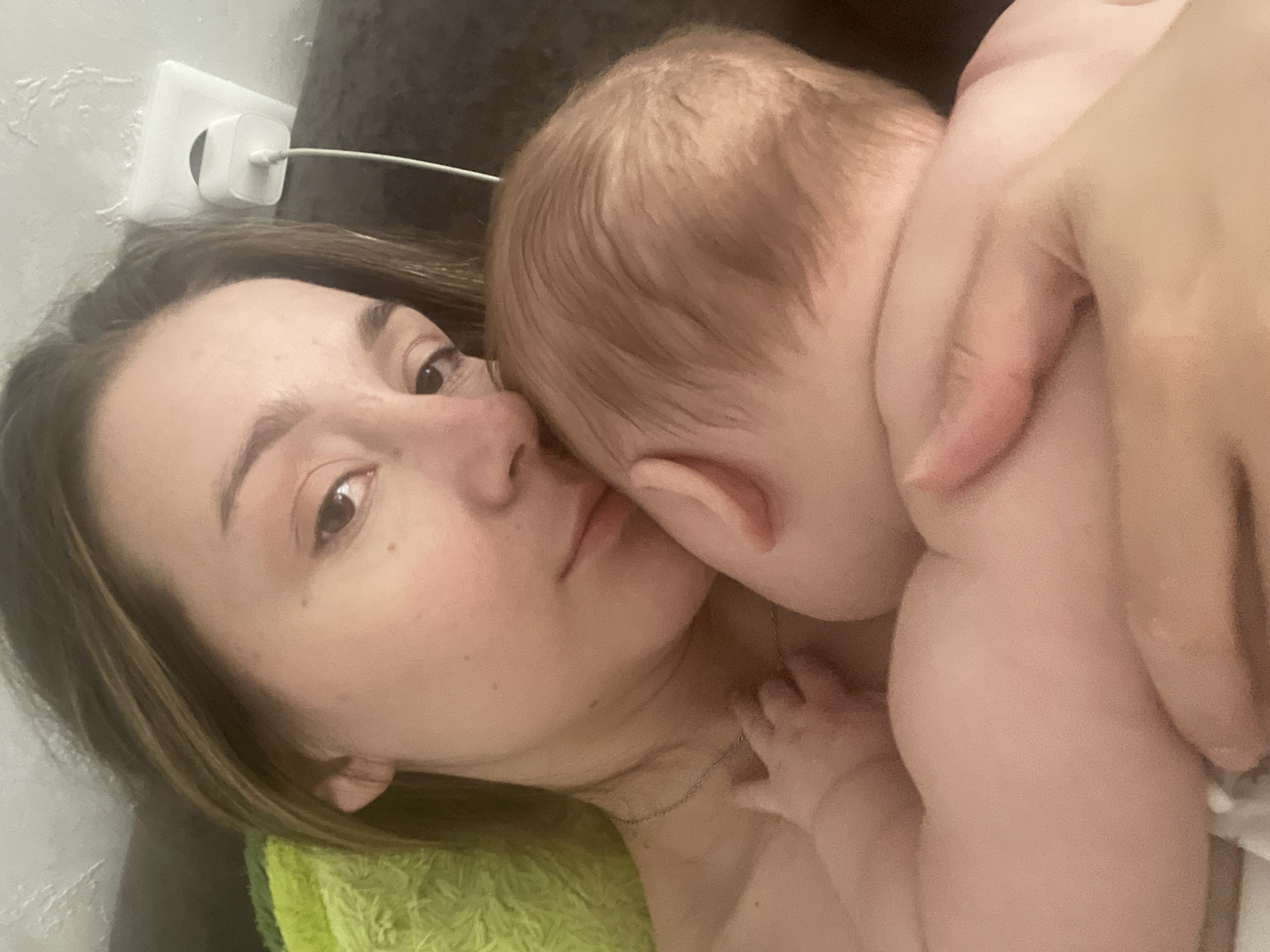 The image is a close-up selfie of a woman holding a baby. The woman has light brown hair and is looking towards the camera with a soft expression. The baby is snuggled against her chest, with its face turned away from the camera. The background is blurred but appears to be an indoor setting with a white electrical outlet on the wall. The image does not provide any information about the location in Stavropol, Russia.