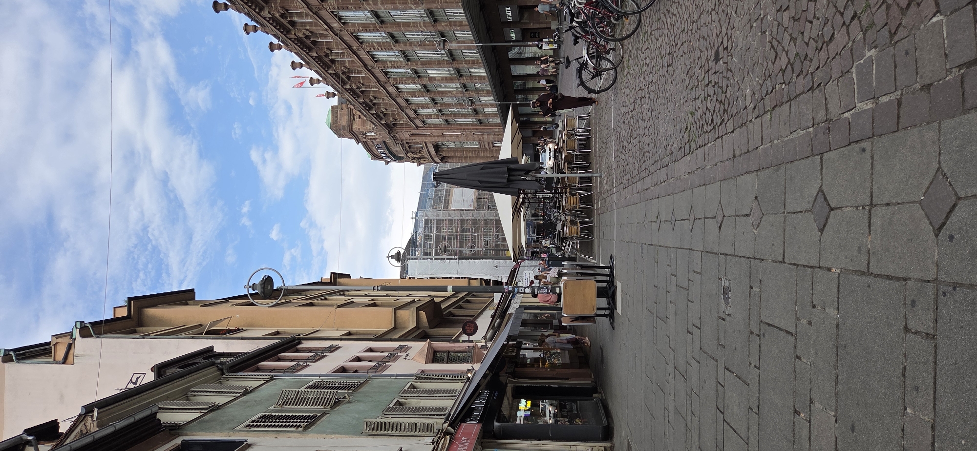 The image depicts a street scene in Strasbourg, France, likely in a commercial or pedestrian area. On the left, buildings with shops on the ground floor line the street. The architecture appears to be a mix of older styles, with shutters on the windows. A street lamp with a circular frame is visible.
The right side of the street features a larger, ornate building with multiple windows and architectural detailing, possibly a department store, with signage that includes "GALERIES LAFAYETTE". In front of this building, an outdoor seating area for a cafe or restaurant is set up with tables and chairs under large umbrellas.
Several bicycles are parked along the right side of the street. A few individuals are walking in the scene, including a person in a brown robe walking on the cobblestone section of the street, and others near the shops and cafe. Construction scaffolding is visible on a building in the background, suggesting ongoing renovation or development. The sky above is partly cloudy. The ground is paved with a combination of large stone slabs and smaller cobblestones.