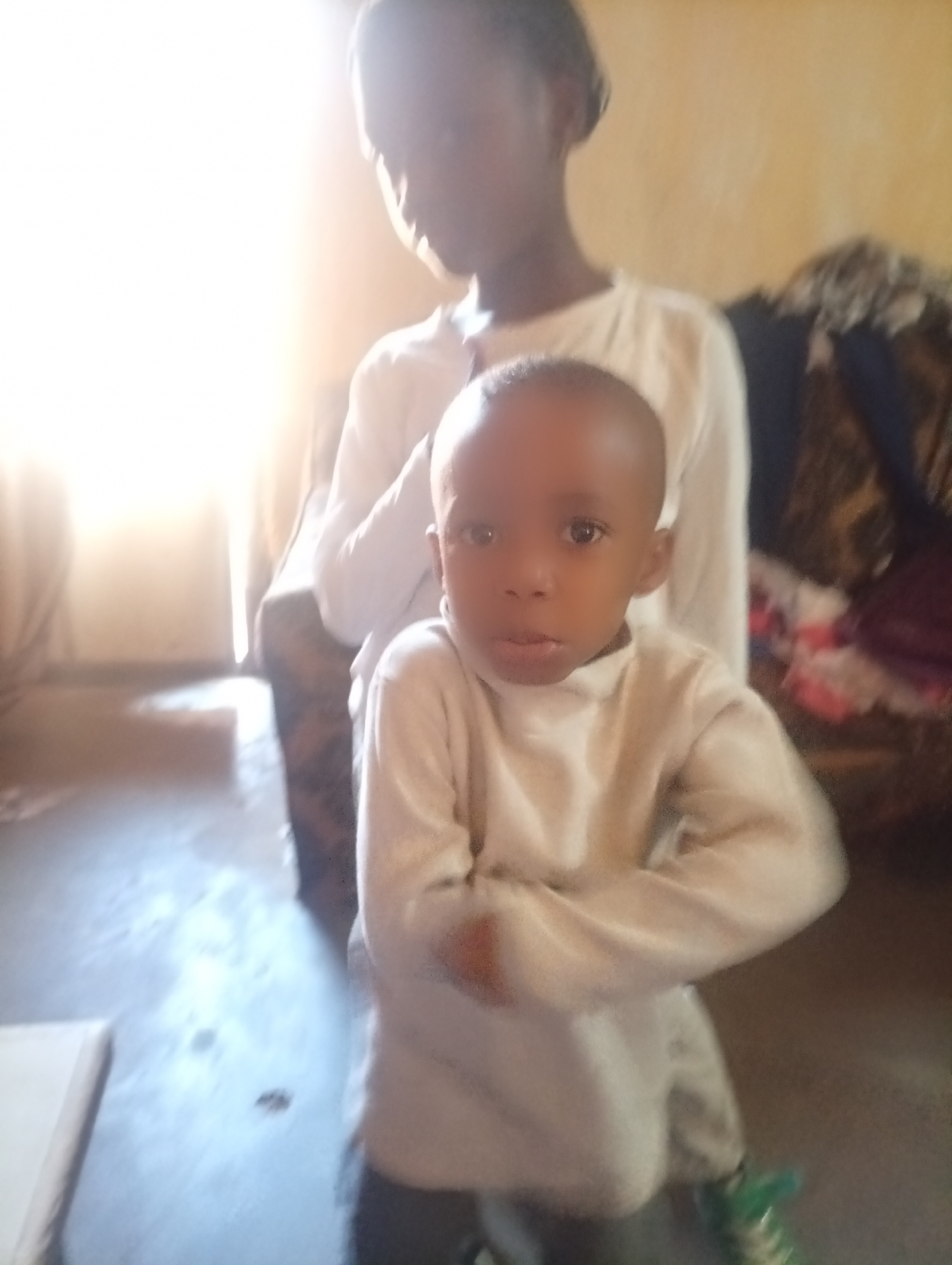 An interior scene in Sabon Tasha, Nigeria, features two individuals of African descent. In the foreground, a young child, approximately 3-5 years old, stands centrally, facing the camera with a neutral expression. The child has short dark hair and is wearing a light-colored (cream or white) long-sleeved turtleneck shirt and dark trousers, with part of a bright green shoe visible on the right foot. His arms are crossed in front of his chest. Behind the child and slightly to the right stands an adult, likely female, whose head and upper torso are visible. She has dark hair, possibly pulled back, and wears a light-colored long-sleeved top and dark lower garments. Her gaze is directed away from the camera, and she is partially out of focus.
The room has light-colored (pale yellow or beige) walls. Strong natural light enters from the left side of the frame, creating an overexposed area and illuminating parts of the dark, possibly concrete or tiled floor. A dark, patterned fabric is visible on the right side of the frame, behind the adult, potentially part of furniture or draped material. A light-colored object, possibly a book or a piece of furniture, is partially visible on the floor in the bottom left corner. The environment appears to be a modest indoor setting.