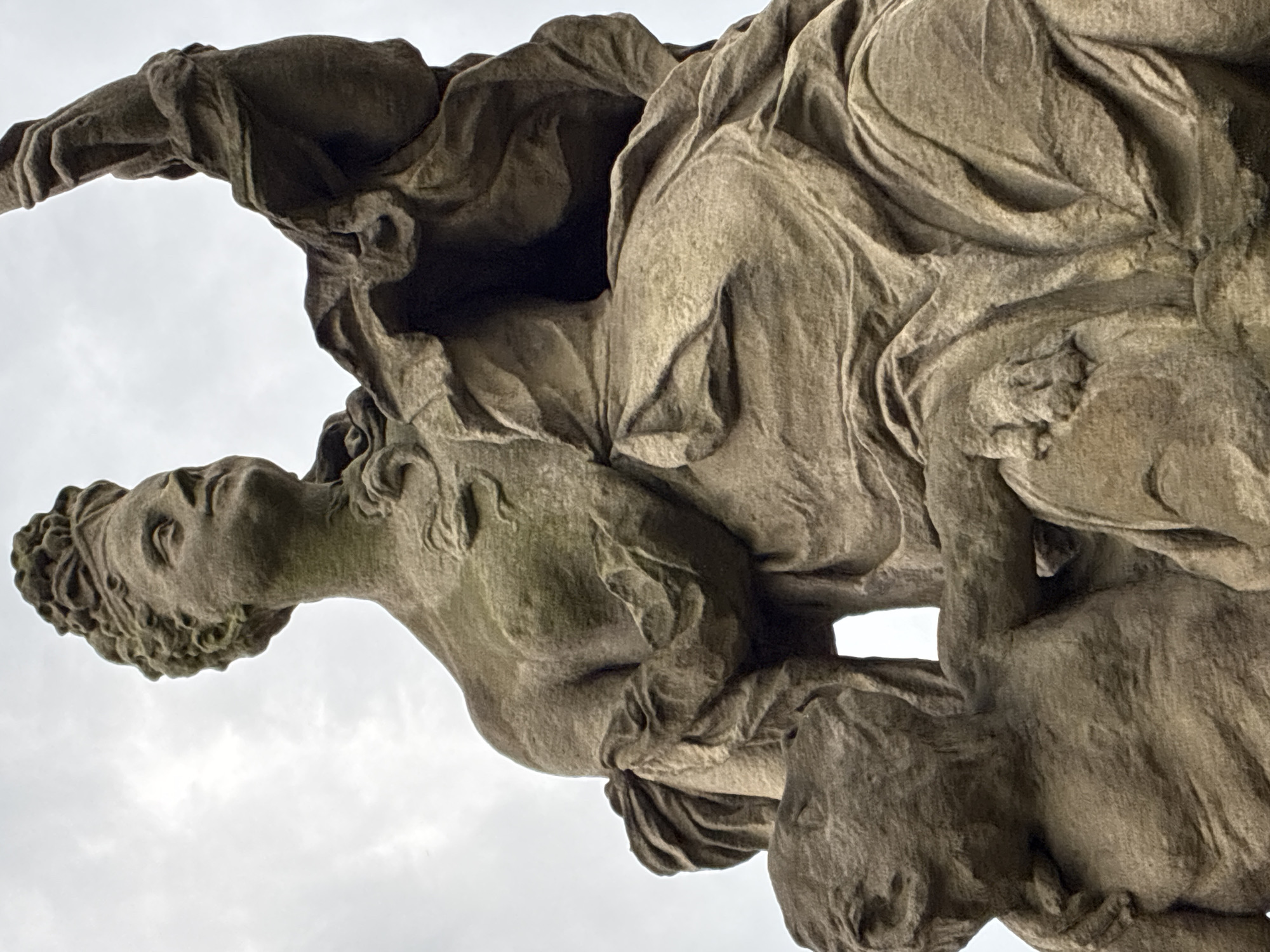 This is a low-angle, close-up photograph of a weathered stone sculpture in the city of Kuks, Czechia. The sculpture depicts a regal female figure in elaborate, draped clothing, holding an object aloft in her right hand. Her head is turned upwards and to the left, with her hair styled in an intricate updo. To her left, a smaller, childlike figure is visible, with its arm around the female figure's body. The sculpture is rendered in a Baroque style, characterized by dramatic movement and detail. The stone is aged, with visible texture and some signs of erosion, suggesting it is an outdoor monument. The background consists of an overcast sky.