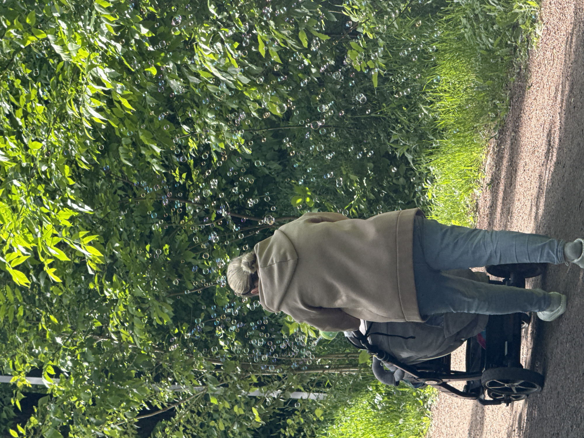 A person wearing a tan hooded sweatshirt and jeans pushes a stroller on a path bordered by lush green vegetation. Bubbles float in the air. The location is likely in a park in Moscow, Russia. The scene depicts a casual outdoor setting, possibly a parent or caregiver enjoying a walk with a child.