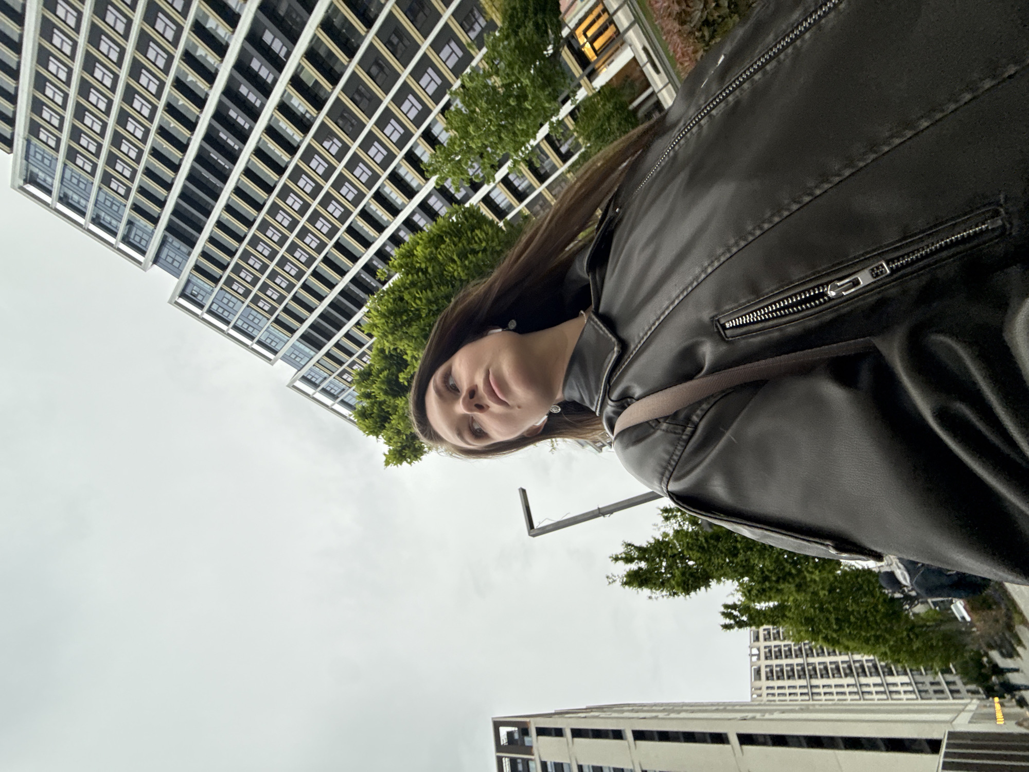 The image, rotated 90 degrees clockwise, presents a low-angle view of a young woman against an urban backdrop under an overcast sky in Belgrade, Serbia.
The primary subject, a woman with long, dark hair, occupies the right side of the frame, gazing downwards towards the camera with a neutral expression. She is wearing a dark, possibly black, leather jacket with prominent metallic zippers on the front and pockets. A light-colored strap, potentially from a shoulder bag, crosses diagonally over her chest. An earring or earbud is visible in her visible ear.
The background features several tall, modern buildings. A large building with a repeating pattern of rectangular windows and dark panels dominates the upper left and central portion of the frame, characterized by its white or light-grey structural outlining. Other urban structures are partially visible in the lower right. Lush green trees are interspersed between the buildings, adding a touch of nature to the cityscape. A dark, angular street fixture, possibly a lamp or signpost, extends into the frame from the mid-right.
The sky is a uniform, bright grey, indicative of an overcast or cloudy day, providing diffused lighting. The overall scene suggests an urban setting. The activity appears to be a casual portrait or selfie taken from a significantly low perspective looking upwards. No visible text is present in the image.