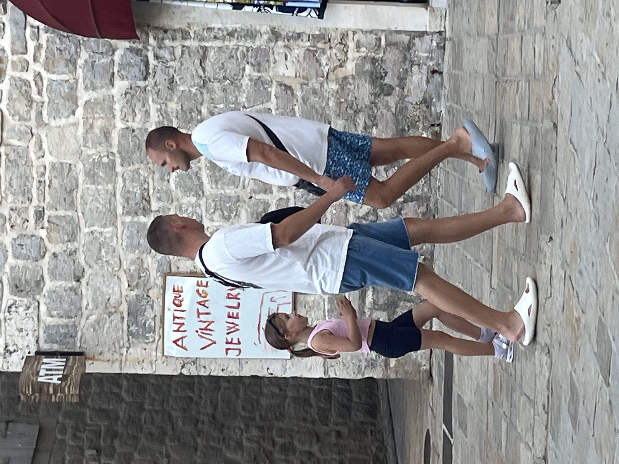The image depicts a street scene in Budva, Montenegro. In the foreground, a young girl with blonde hair in a ponytail, wearing a pink tank top and black shorts, walks on a cobblestone path. She is accompanied by two men, one on her left and one on her right. The man on her left, wearing a white t-shirt and blue denim shorts, is looking at the girl. The man on her right, also in a white t-shirt and blue patterned shorts, is looking away from the girl, towards the left side of the image. A sign reading "ATM" is visible on a stone building to the left, and another sign below it says "ANTIQUE VINTAGE JEWELRY". The background consists of a stone wall and a reddish awning. The overall lighting suggests it is daytime.