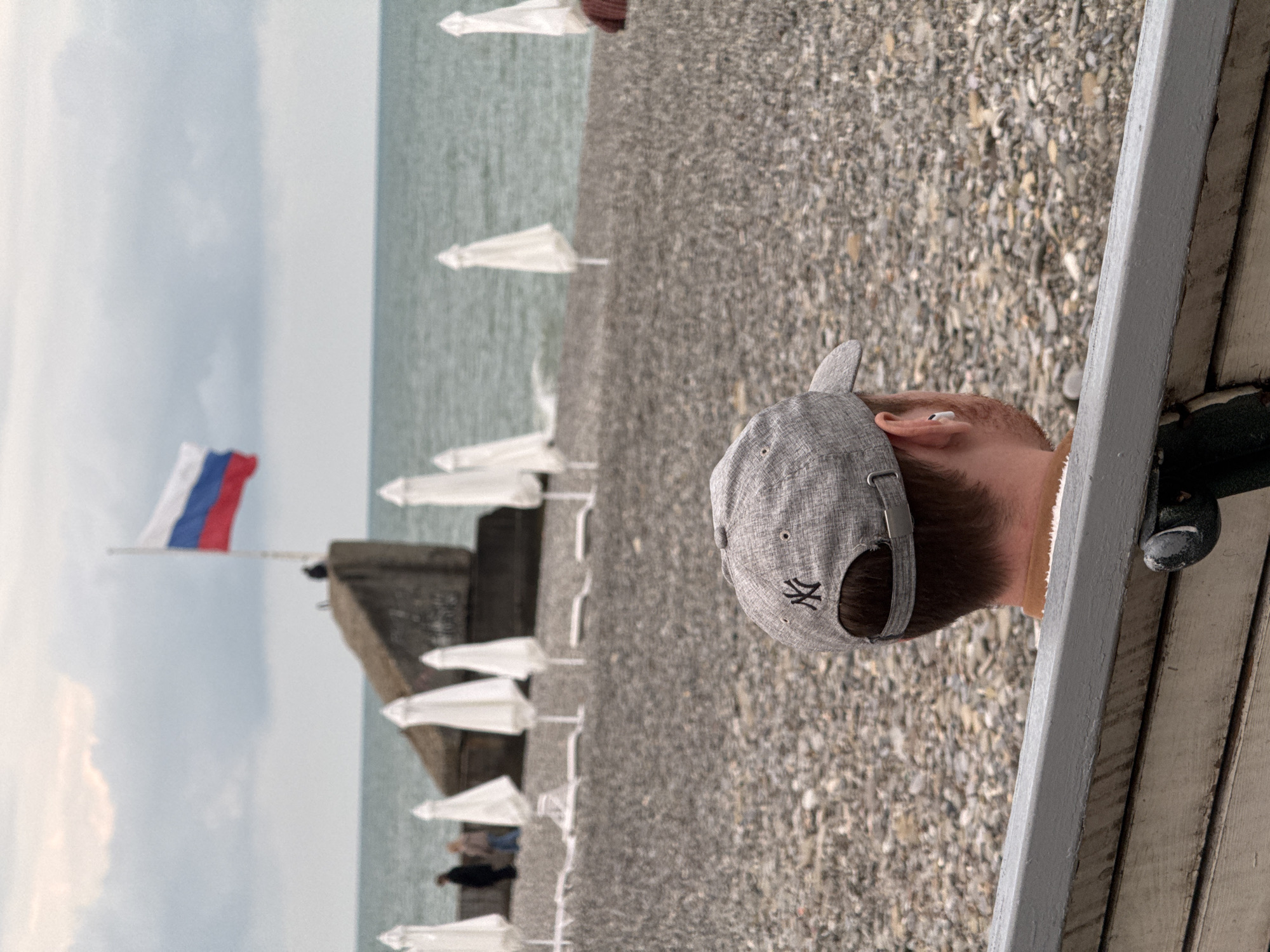 A man wearing a gray baseball cap is sitting on a wooden bench at a rocky beach on a cloudy day in Sochi, Russia. The beach is lined with closed white umbrellas and has a Russian flag on a pole in the distance. The water is visible beyond the beach.