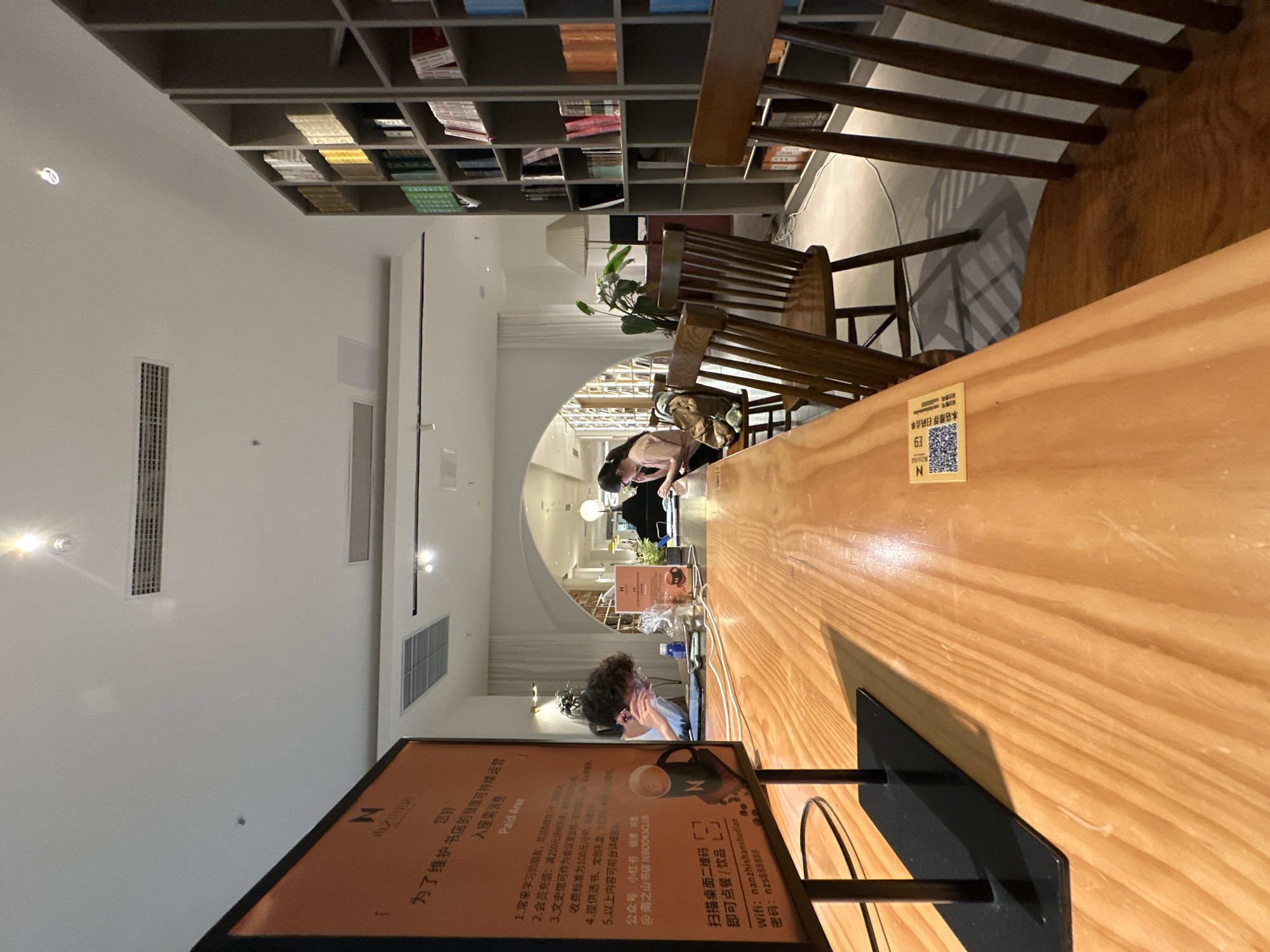 The image depicts the interior of a coffee shop or bookstore in Chongqing High-tech Zone, China. The scene is viewed from a low angle, focusing on a wooden table in the foreground. A large orange sign with Chinese text is on the left, partially obscured by the table. The sign appears to be a menu or information board.
On the table, a person with curly hair is seated and looking at a tablet. To their right, another person is seated at a computer. In the background, there are bookshelves filled with books, a plant, and more tables and chairs. The overall lighting is warm and ambient, suggesting it might be evening or indoors. The environment appears to be modern and well-lit, with a mix of seating arrangements for patrons. The wooden furniture and bookshelves contribute to a cozy and inviting atmosphere.