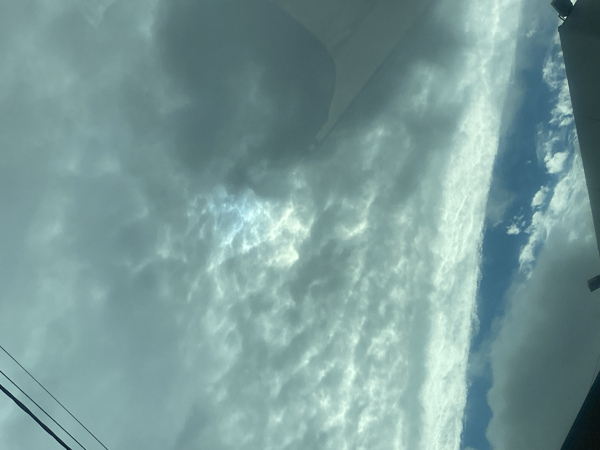 The image shows a cloudy sky with some breaks in the clouds where sunlight is visible. There are also some dark clouds and what appears to be a power line or cable in the upper left corner. The lower part of the image shows a dark silhouette of structures, possibly buildings or vehicles, against the sky. The scene appears to be outdoors.