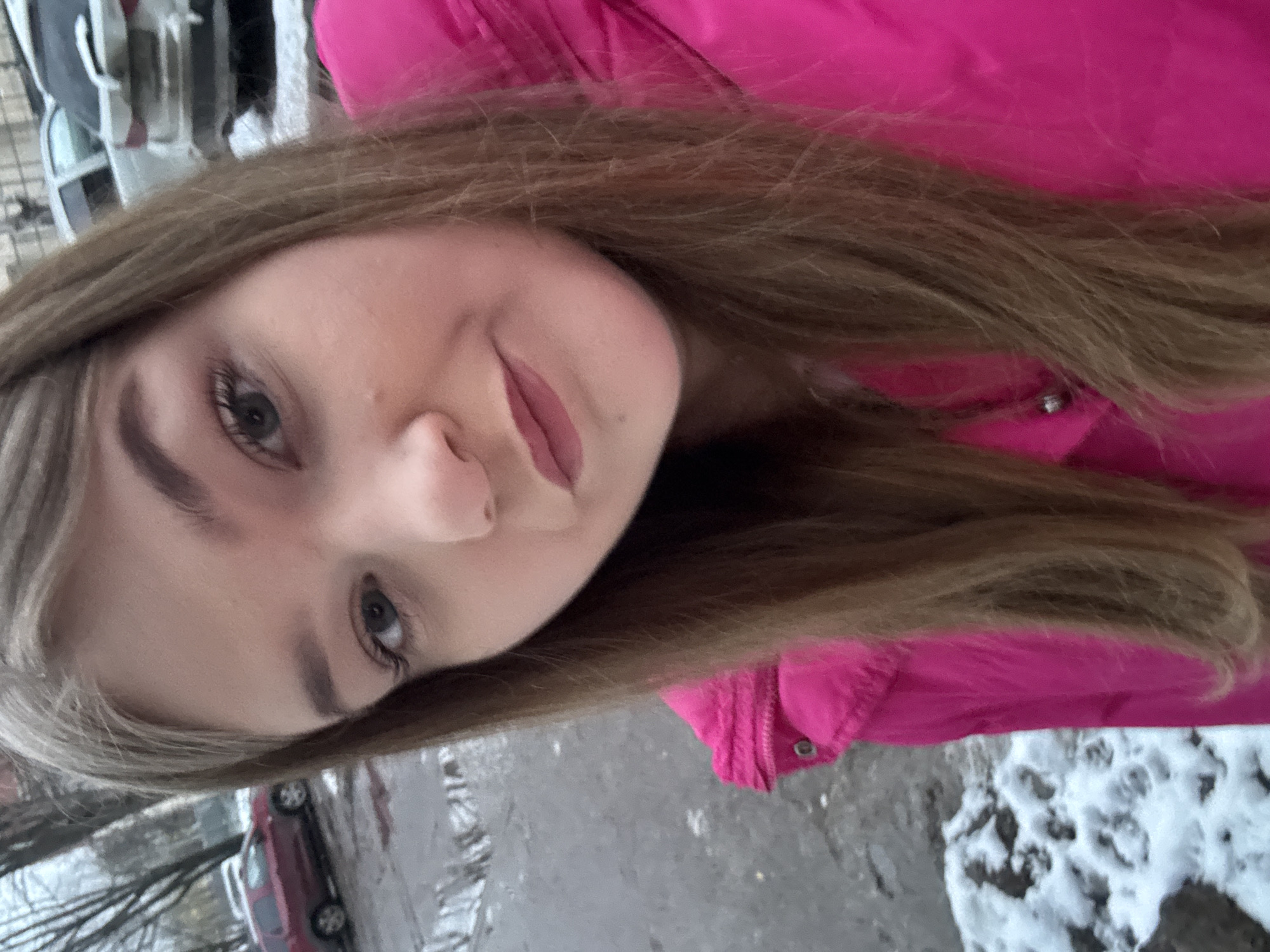 A young woman is posing for a selfie outdoors. She has light brown hair and is wearing a bright pink jacket. The background shows a snowy street scene, likely in the city of Chaikovsky, Russia, given the prompt. There is a road with patches of snow, trees, and parked cars visible.