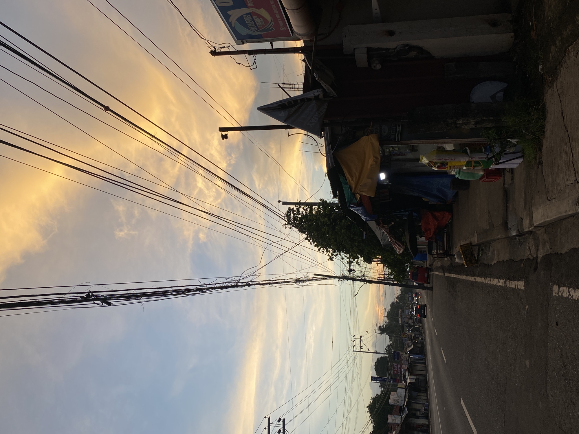 A street scene in Davao City, Philippines, captures the transition from day to night with a vibrant sunset sky. The image features a two-lane asphalt road with white lane markings, flanked by a sidewalk lined with small shops and stalls. Overhead, a complex network of electrical wires crisscrosses the frame, supported by utility poles. The shops on the right side of the road are constructed with corrugated metal and tarpaulin coverings, displaying various goods and signs, including one for "Robbus Printing Press". On the left, a row of smaller establishments are visible, some with signage. Several vehicles, including cars and a red van, are on the road. The sky is painted with hues of orange, yellow, and blue, indicating the sunset. The overall impression is of a bustling urban environment under a dramatic sky.