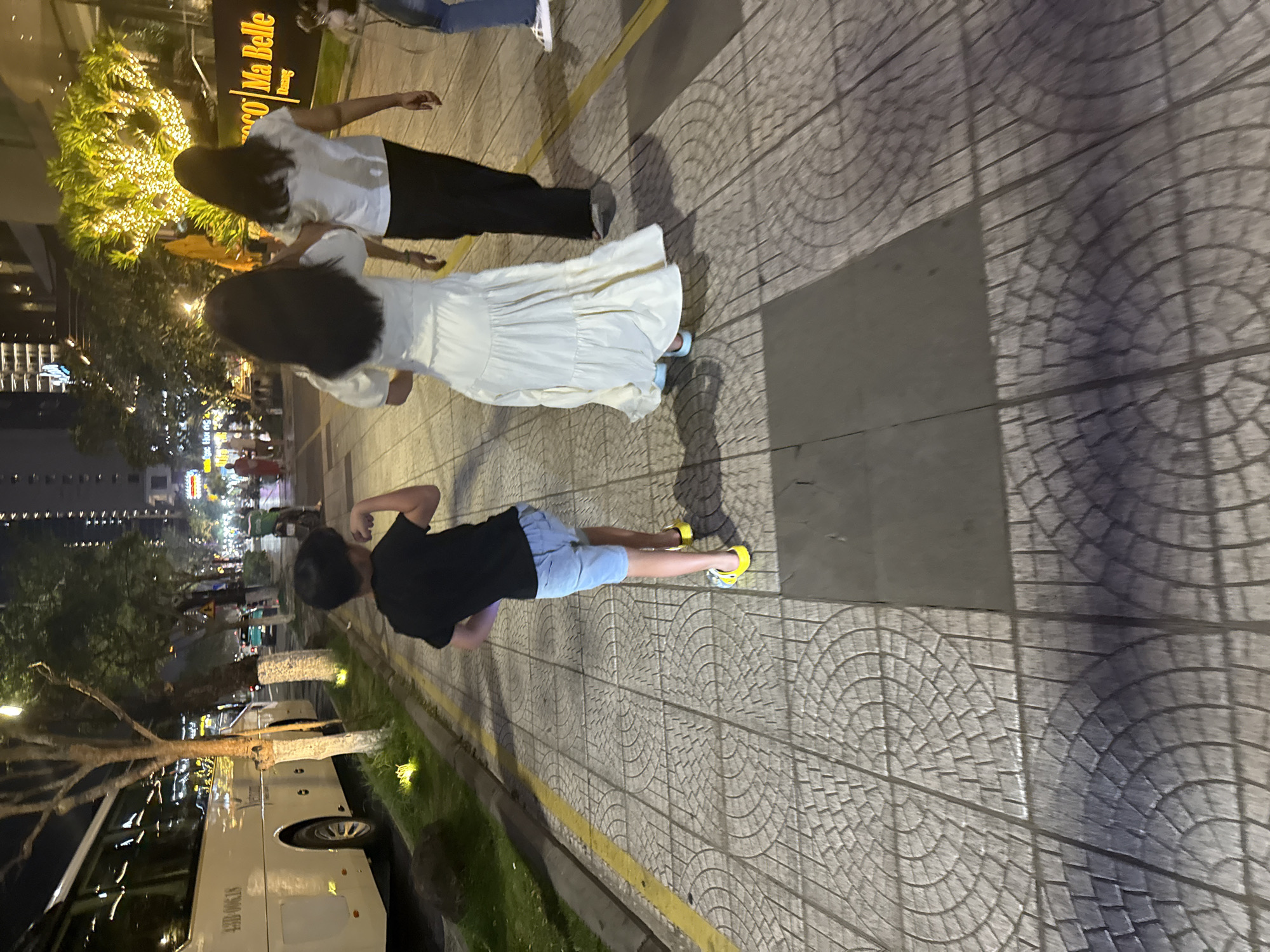 The photograph, taken at night, shows a street scene in Đà Nẵng, Vietnam. Pavement with a circular pattern makes up the foreground. A child in a black shirt, blue shorts, and yellow shoes stands on the pavement. Two women with long dark hair stand nearby, one wearing a white top and black pants, the other in a long white dress. A large white bus is parked along the curb on the left side of the image. In the background, lights illuminate buildings and trees along the street, including a sign for "Marco Ma Belle" over a shrub or tree covered in lights. The sky is dark.