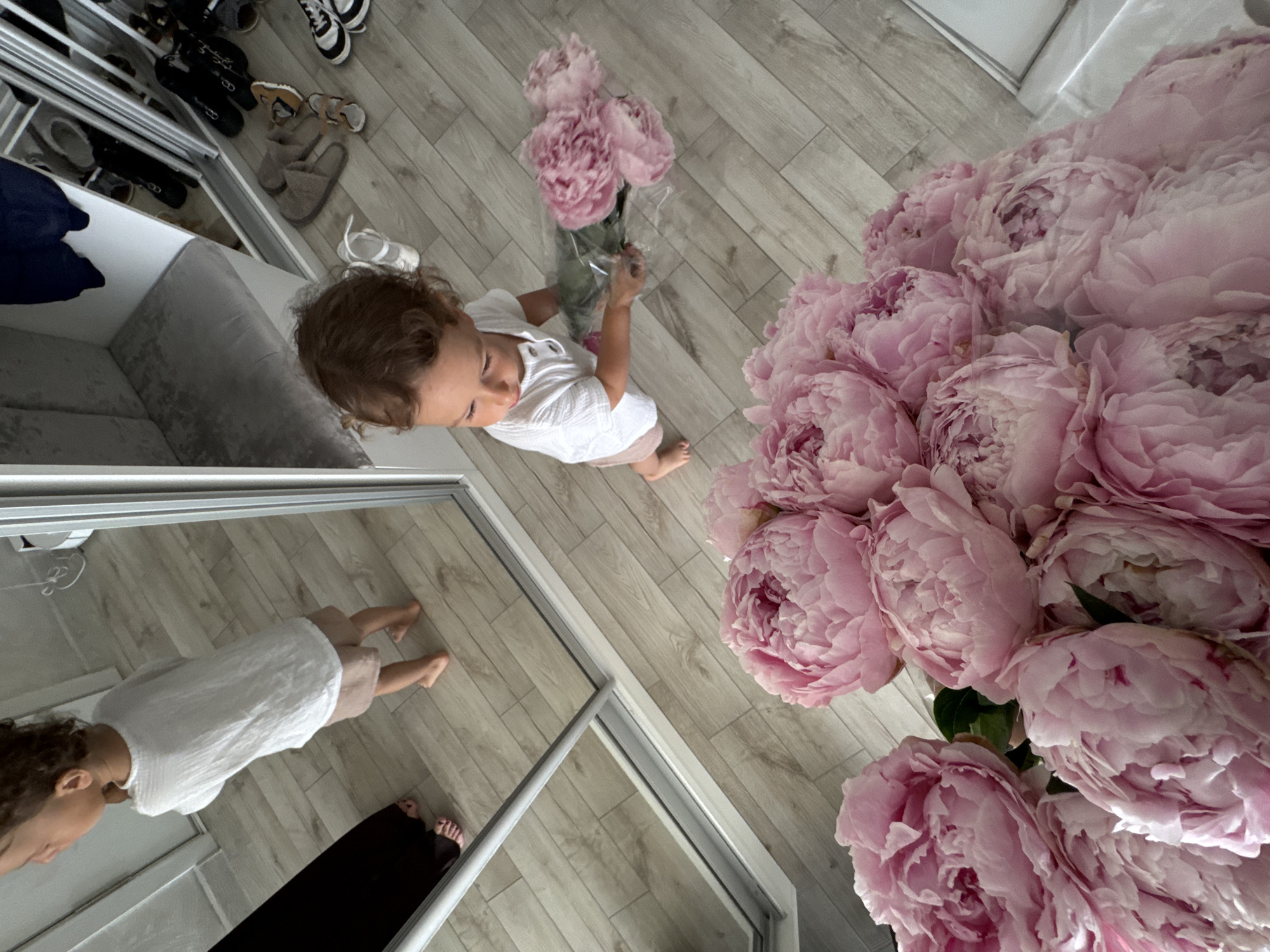 An overhead shot captures a young child holding a bouquet of pink peonies, positioned in the foreground. The child, dressed in a white shirt and light brown shorts, has curly brown hair and is looking down at the flowers, which are partially wrapped in clear plastic. The background reveals a room with light wood-paneled flooring and a large mirrored wardrobe. In the reflection of the wardrobe, another young child wearing a white t-shirt and brown shorts is visible from the back, standing near a black garment. A bench with grey upholstery is also reflected in the mirror, along with various shoes and slippers scattered on the floor in front of the wardrobe. The overall scene suggests an indoor setting, possibly a residential interior, given the presence of personal items and a wardrobe. The lighting is bright, illuminating the details of the flowers and the environment. There is no indication of the city of Sochi, Russia, within the image content itself.