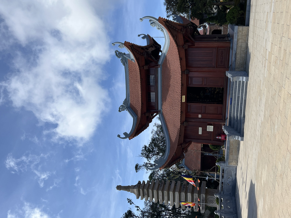 The image displays an outdoor architectural complex, identified as a Buddhist pagoda or temple, located in an unknown city in Vietnam. The primary structure is a two-tiered building constructed from reddish-brown wood, featuring red terracotta tiled roofs with distinctively curved, ornate grey-blue eaves adorned with decorative elements. A central entrance, comprising partially open double wooden doors, leads into a dark interior. A flight of grey stone steps ascends to this entrance from a large, light-colored paved plaza, made of square tiles. To the left of the main building, a tall, multi-tiered stone stupa or pagoda stands, light grey in color, with colorful Buddhist flags (yellow, red, blue, white, orange) visible at its base. A low grey stone balustrade runs along the edge of the plaza near the stupa. The background reveals dense green foliage from trees, suggesting a natural or elevated setting. The sky is bright blue with scattered white cumulus clouds. No individuals, specific actions, or interactions are visible within the frame.