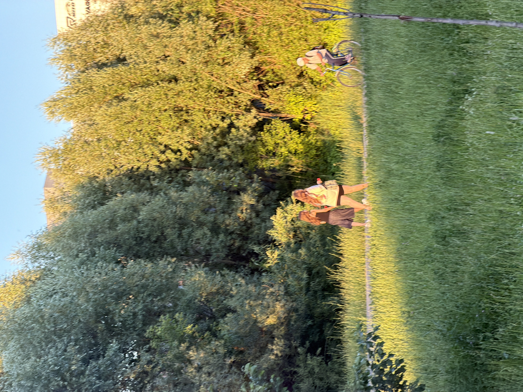 A wide shot captures a park scene with a paved path winding through lush green grass. On the path, two young women walk together, one in a dark skirt and top, the other in a light-colored top and shorts. To the right, a person on a bicycle, wearing a light-colored shirt and dark pants, rides away from the camera. The background is filled with dense green trees, bathed in the warm glow of the sun, with a building visible beyond the foliage on the right. The sky is a clear blue.