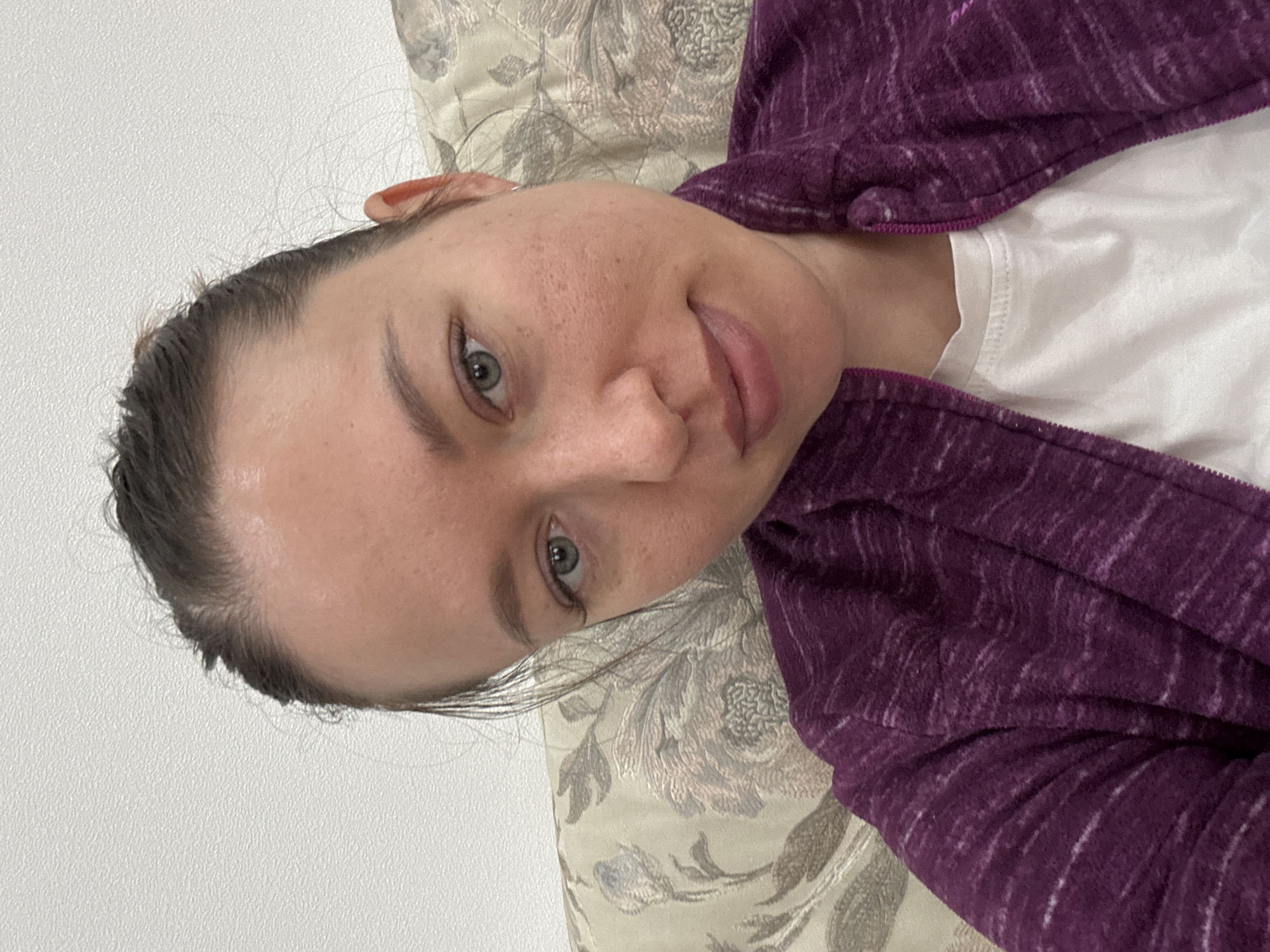 A woman with fair skin, grey eyes, and dark brown hair pulled back from her forehead is lying on a pillow with a floral pattern. She is wearing a white t-shirt and a purple zip-up fleece jacket. The background is a white wall. The location is Ферма, Russia, but there's no further information to deduce the setting or details about the city.