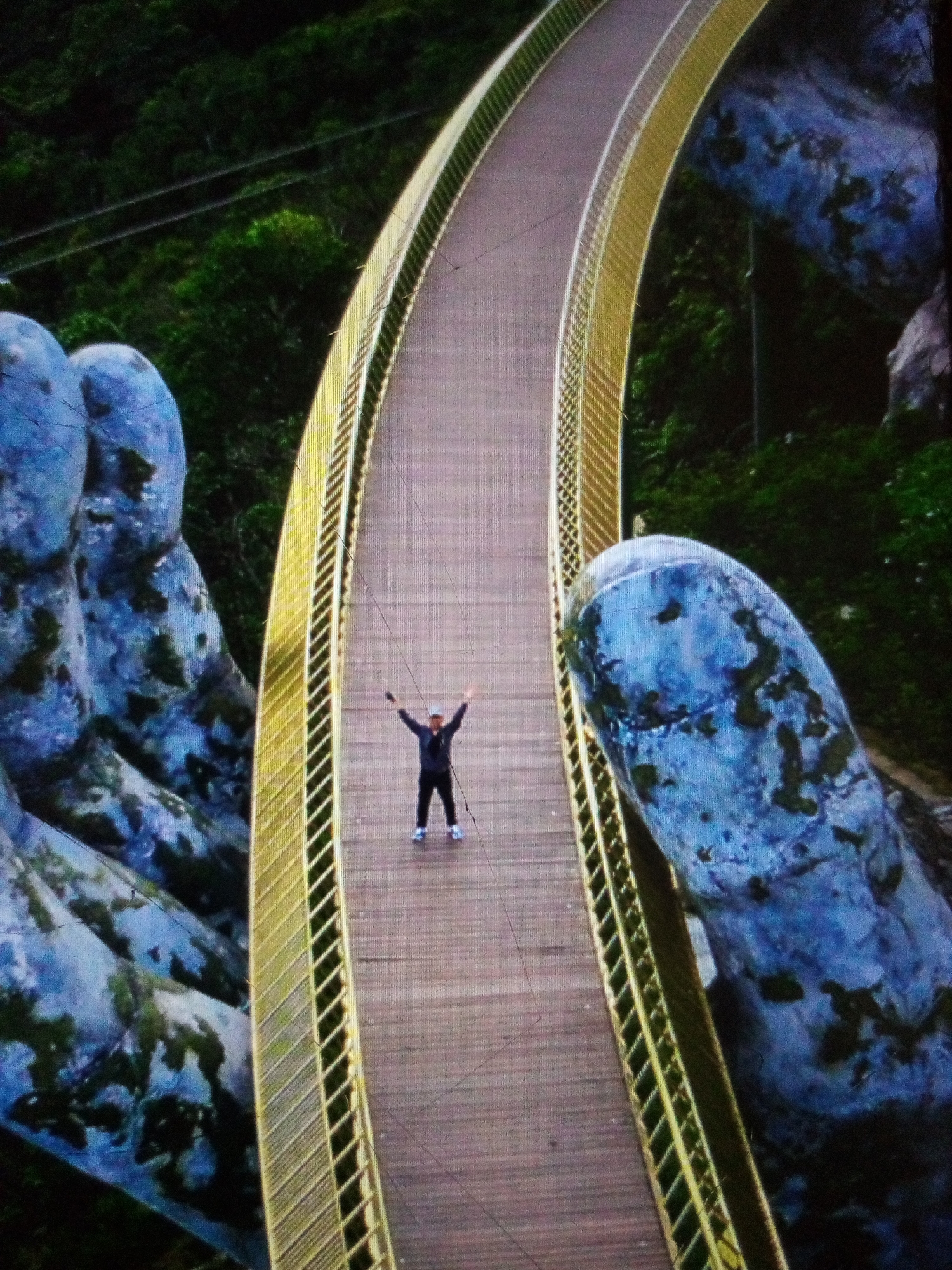 The image shows a person standing in the middle of a wooden bridge with a golden railing. The person is wearing a dark jacket and pants and has their arms raised in the air. The bridge is curved and appears to be elevated, with lush green trees visible in the background. On either side of the bridge, there are large, blue, stone-like structures that resemble giant hands. The scene is an aerial view, looking down on the bridge and its surroundings. The location is the Golden Bridge in Da Nang, Vietnam, not Astana, Kazakhstan.