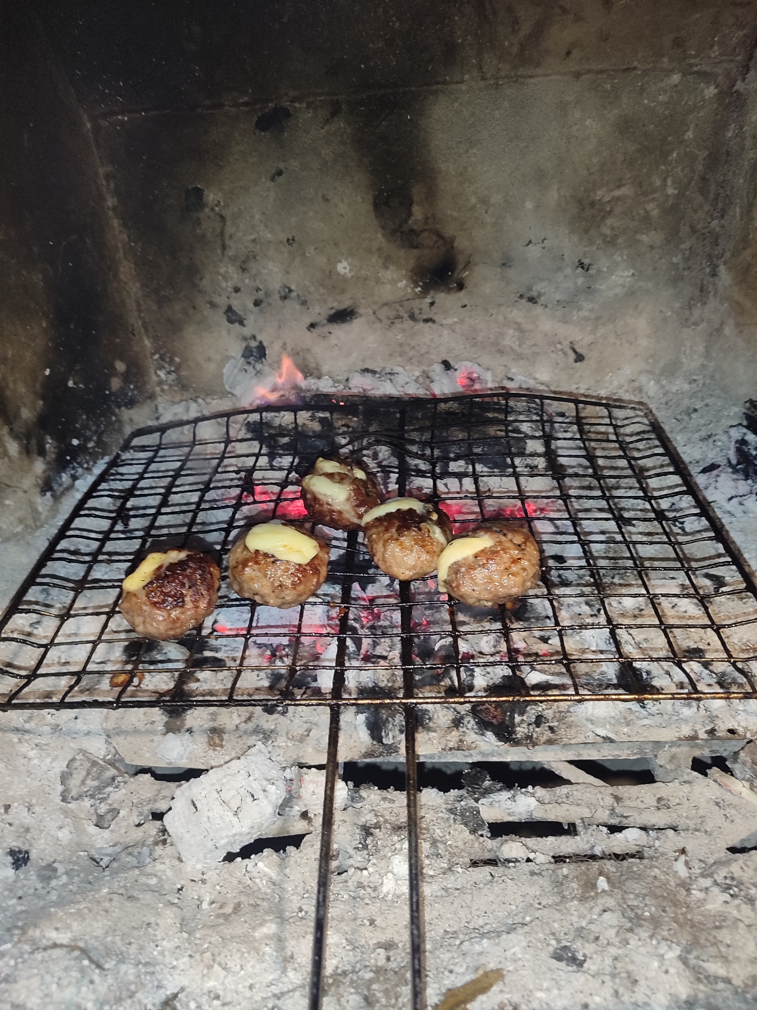 The image depicts a rudimentary indoor grill setup, possibly within a fireplace. On a metal wire grill, four cooked, round patties are placed, each topped with a yellowish substance that appears to be melting cheese. Below the grill are embers and ash, suggesting an active or recently active fire. The surrounding structure appears to be made of stone or concrete and is heavily stained with soot and residue, giving the impression of a rustic and utilitarian cooking environment. While the location of Varshilo, Bulgaria, is mentioned, no details within the image confirm this information. There are no people present in the picture, and the primary action depicted is the cooking of the patties.