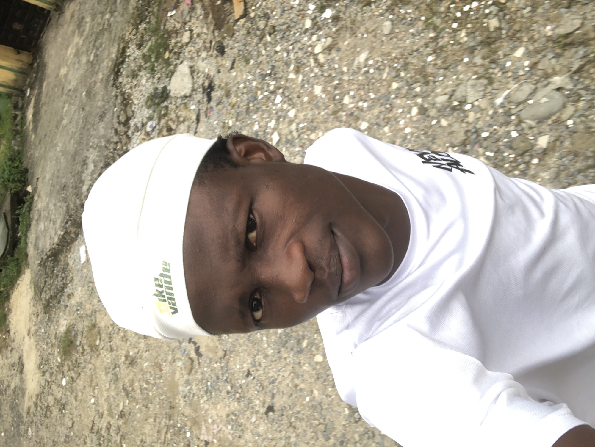 A young man wearing a white t-shirt and a white cap is pictured in an outdoor setting. The cap has a green and yellow logo with the text "Vake Vande". The t-shirt has a black graphic on the left side of the chest. He is looking towards the camera with a slight smile. The background appears to be a dirt and gravel area with some vegetation and a building in the distance. The scene is likely located in the city of Ado, Nigeria, given the context provided.