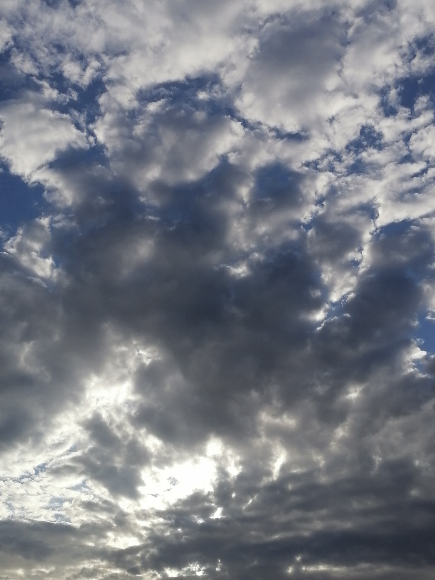 A low-angle, full-frame, medium shot captures a sky filled with a mix of dark gray and bright white clouds. The clouds are textured and appear to be in motion, with sunlight breaking through in some areas, creating a dramatic contrast between light and shadow. The image was taken in Unterhaching, Germany.