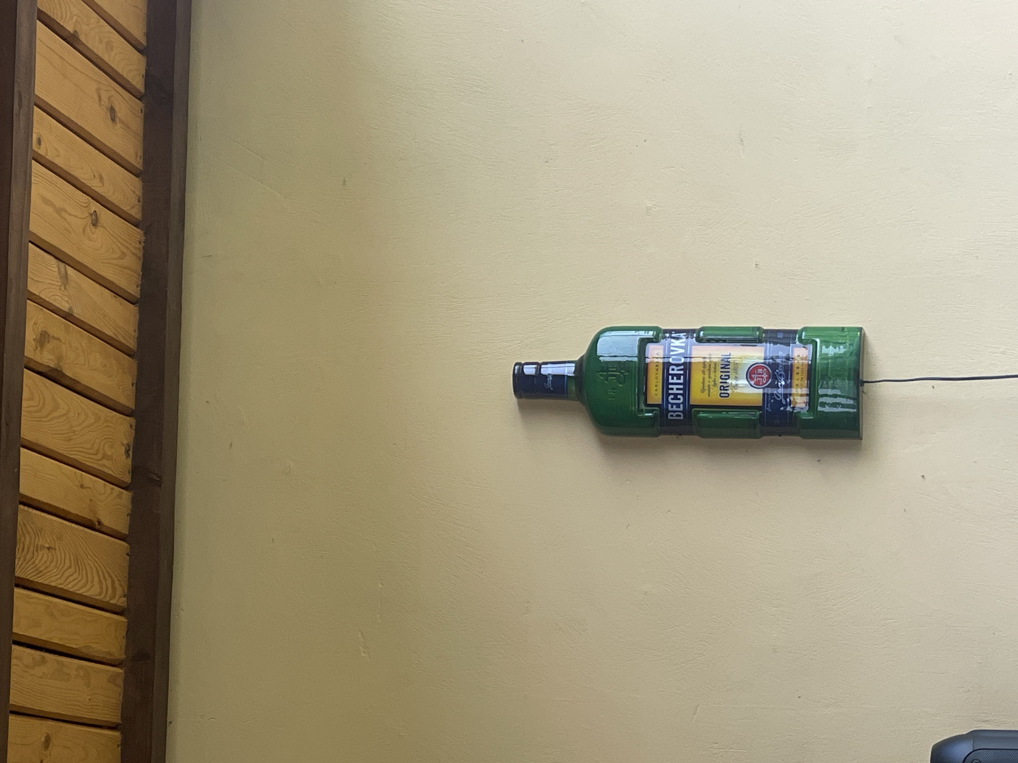 The image depicts a light yellow-beige textured wall in Petrivka, Ukraine, beneath a wooden ceiling structure. The ceiling comprises multiple parallel light-toned wooden planks, secured above a dark brown horizontal beam. Centered vertically on the wall is a large, wall-mounted, green replica of a Becherovka liqueur bottle. The bottle features a blue neck label inscribed with "JAN BECHER" and a main green body label displaying "KARLOVARSKA BECHEROVKA ORIGINAL" along with "Since 1807" and a red circular emblem containing "JB". A thin black electrical wire extends downwards from the base of the bottle replica. No individuals are present in the scene.