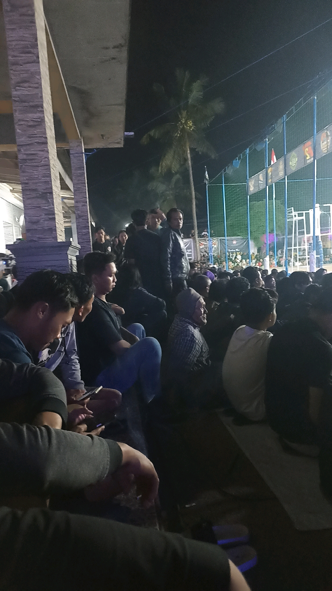 A nighttime exterior scene captures a crowd of people seated and standing in what appears to be a viewing area adjacent to a sports field or event space. The foreground is dominated by the backs and sides of several individuals, some holding mobile phones. In the mid-ground, more people are visible, seated on what looks like tiered seating or the ground. A man in a checkered shirt and head covering is prominent on the right side of the mid-ground. The background reveals a brightly lit net enclosure, likely a sports field, with spectators and possibly event infrastructure. A palm tree is visible against the dark night sky. The setting suggests a public event taking place outdoors in Pacitan, Indonesia, possibly a sports match or cultural gathering, with the architecture on the left suggesting a building structure integrated with the viewing area.