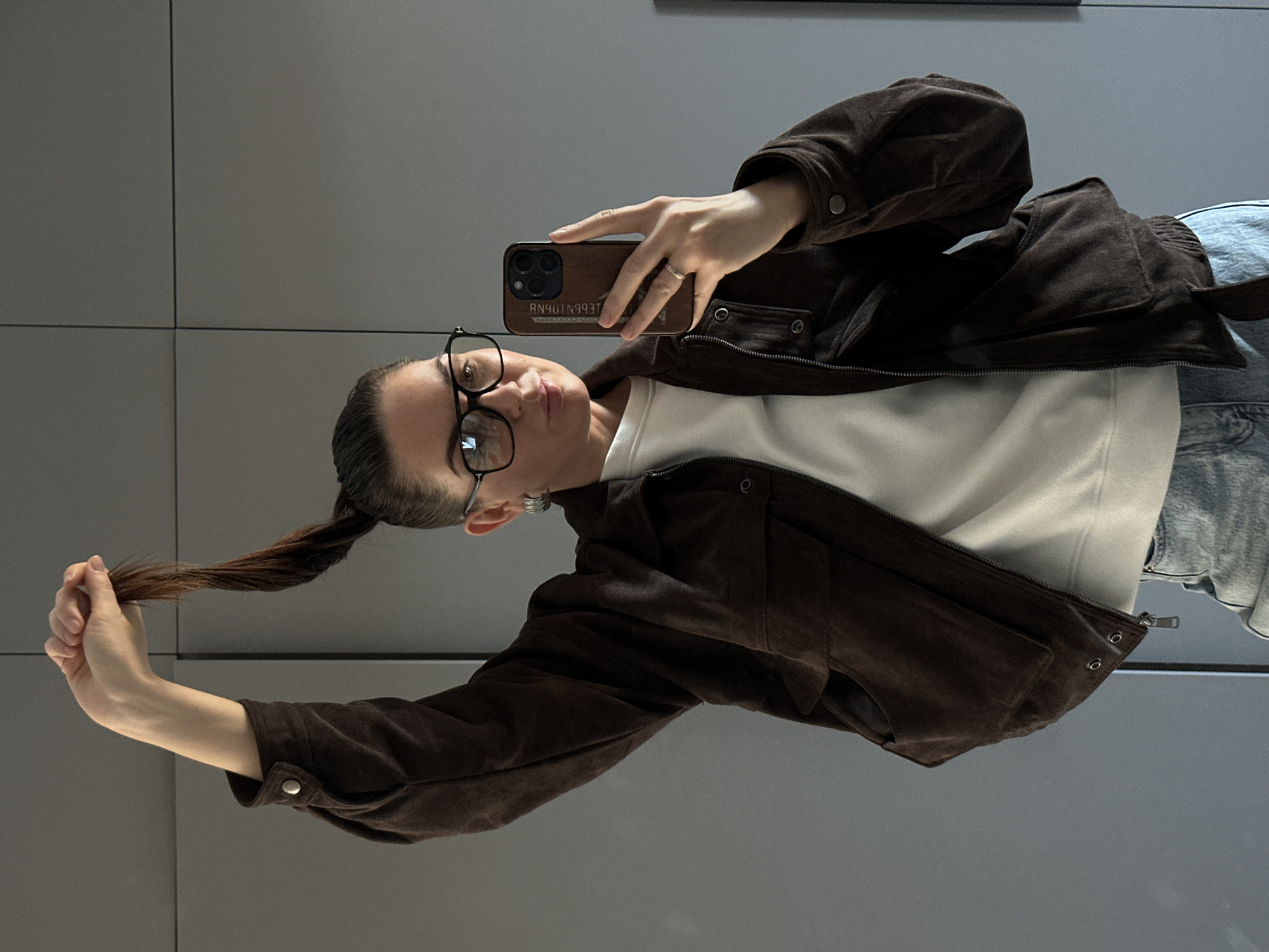 A woman with brown hair tied back in a ponytail is taking a selfie with a smartphone. She is wearing glasses, a white shirt, a brown jacket, and blue jeans. She is holding her ponytail in her left hand. The background is a gray wall. The text on the back of the phone is backwards. The image does not contain any specific details that would allow identification of Chaikovsky, Russia.
