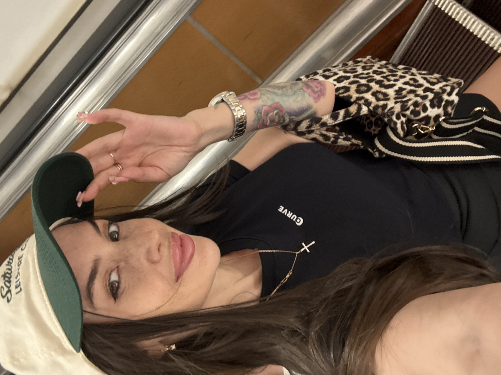 A woman with long dark hair and light skin, wearing a tan and green baseball cap, a black t-shirt with a gold cross necklace, and a leopard print handbag, poses for a selfie on an escalator in St. Petersburg, Russia. The woman has freckles and is wearing makeup, including eyeliner and lip gloss. She has a tattoo of roses on her left arm and is wearing a watch. The background shows the interior of an escalator with brown treads and silver handrails.