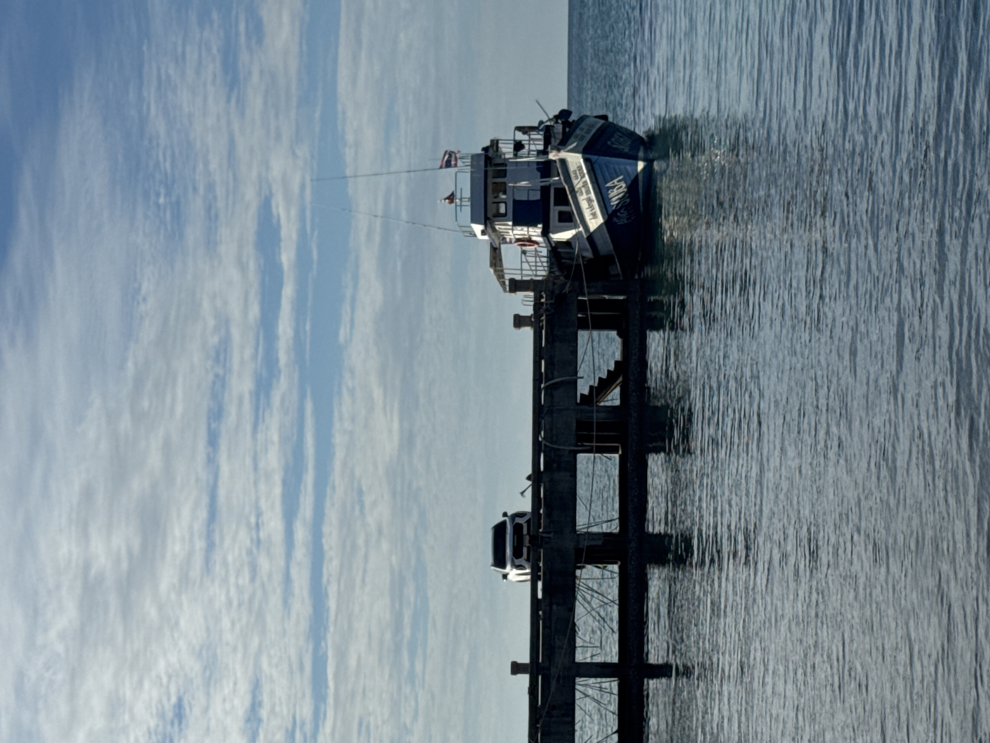 A white SUV is parked on a concrete pier. A boat is docked at the end of the pier. The boat is blue and white with the word "SCUBA" written on its side. The water is calm and the sky is blue with some clouds. The image appears to have been taken in a coastal area of ตำบลเกาะพะงัน, Thailand.