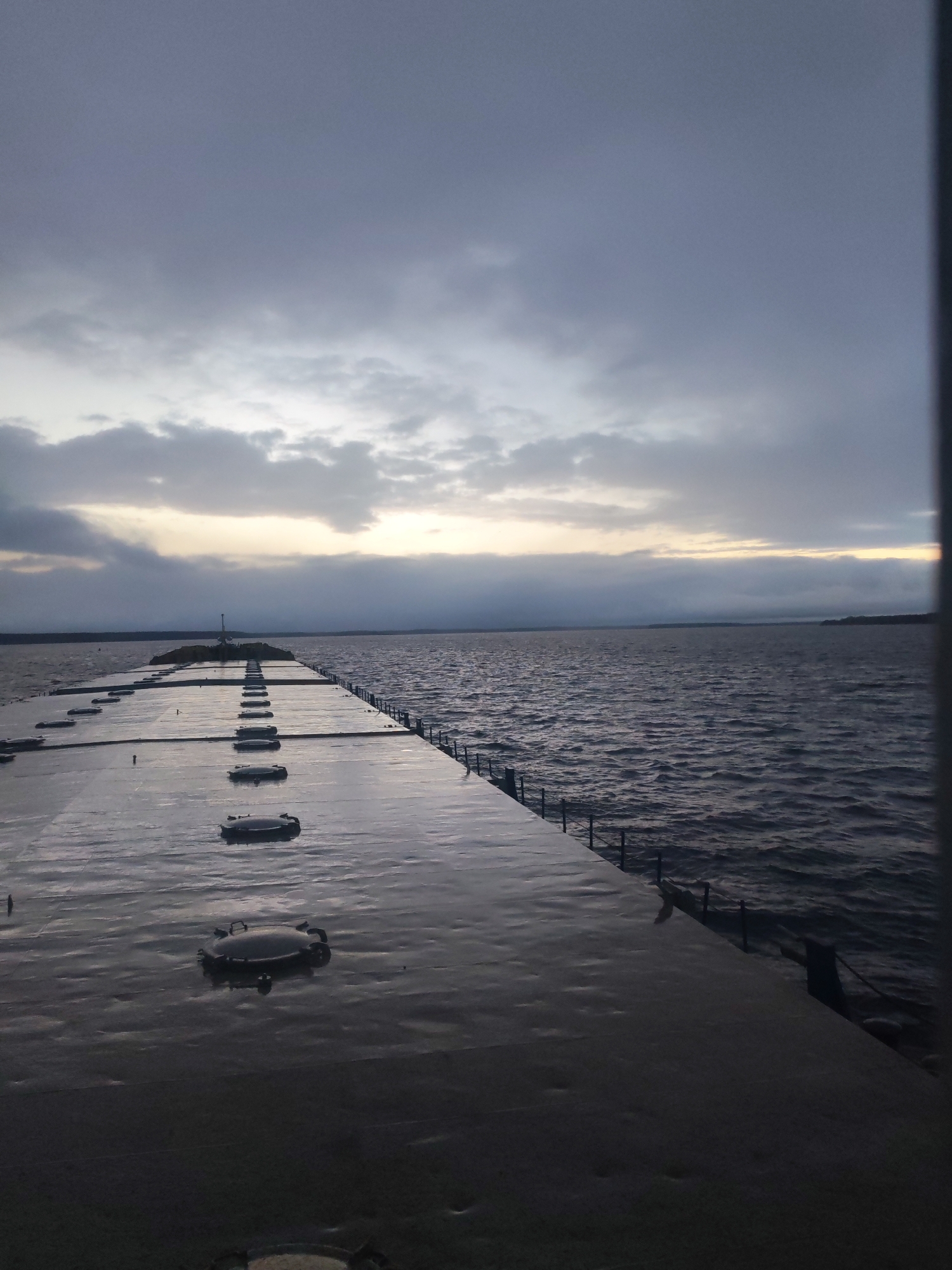 This image depicts a long, grey concrete pier or breakwater extending into a vast body of water, likely a lake or sea. The pier's surface appears wet and reflective, possibly from rain or moisture, and is embedded with numerous evenly spaced, round, dark hatch-like covers. Along the right side of the pier, railings or bollards are visible. At the distant end of the pier, a small structure resembling a beacon or light fixture with an antenna is present.
The water is choppy with visible waves, indicating windy conditions. In the far distance, a faint landmass or coastline stretches across the horizon. The sky is predominantly overcast with heavy grey clouds, but a brighter strip of light near the horizon suggests either early morning dawn or late evening dusk. The overall atmosphere is quiet and somewhat dramatic due to the cloudy sky and vastness of the water. There are no people visible in the image, and no specific activity or event is taking place beyond the natural scene. No text is visible.
