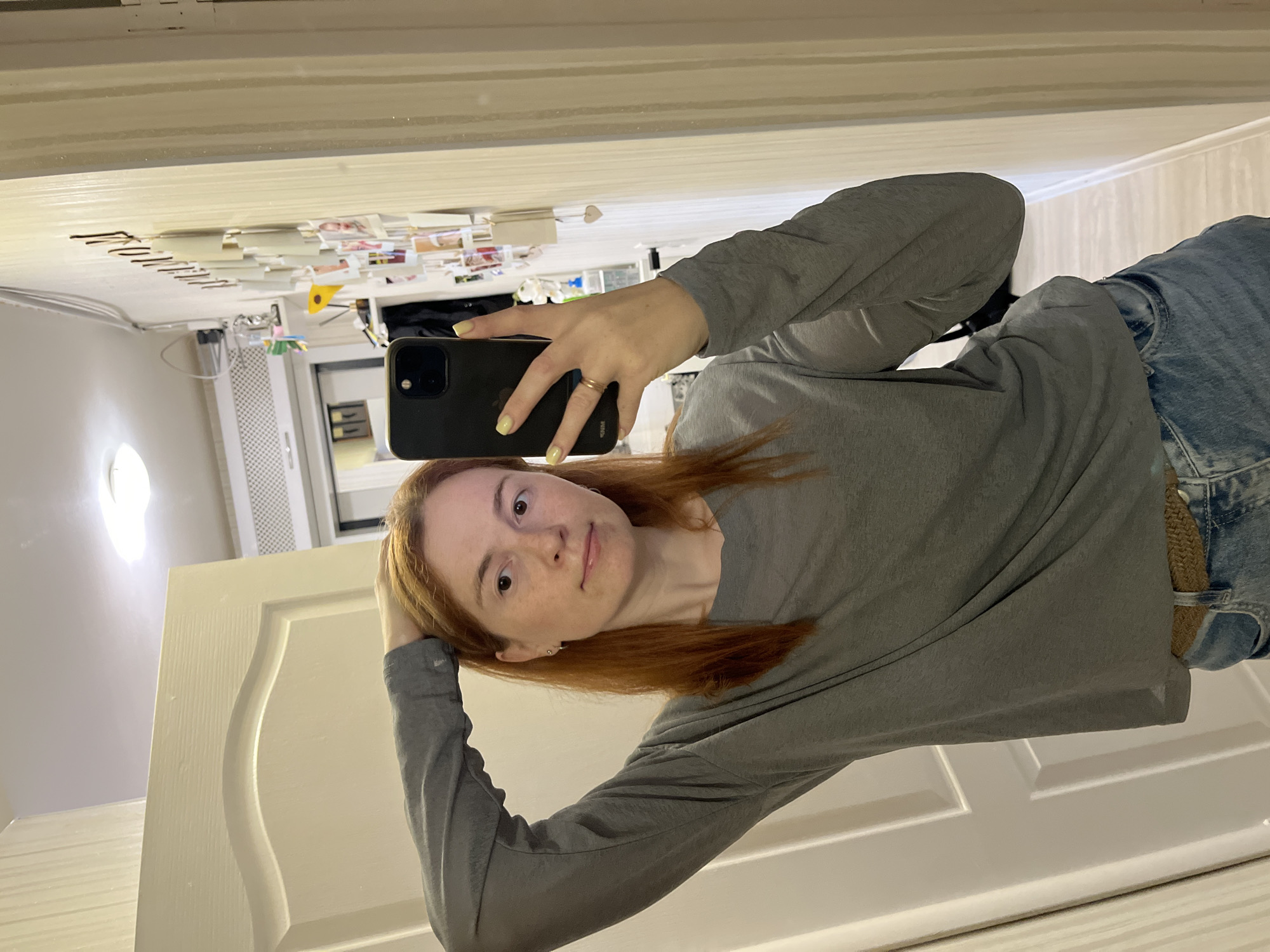 A person is taking a selfie in front of a mirror. The person has red hair, freckles, and is wearing a gray long-sleeved shirt and blue jeans. The person is holding a black smartphone with yellow nail polish. The mirror reflects a room with a white door, a radiator, and a wall covered in photos and other items. The location is potentially Chaikovsky, Russia.