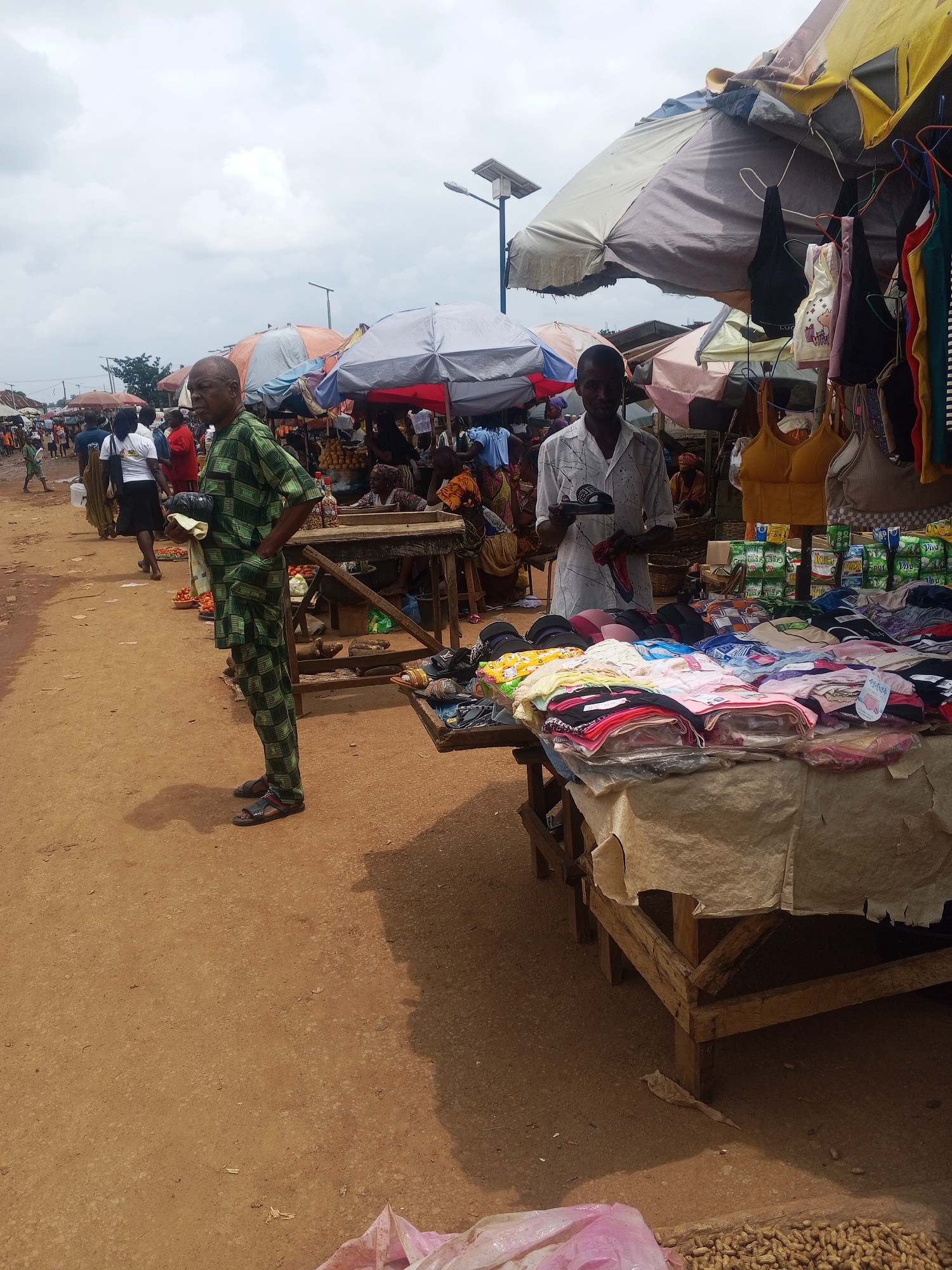 The image depicts a bustling outdoor market scene in Kabba, Nigeria, under a cloudy sky. In the foreground, a man dressed in a green and black patterned traditional African outfit, stands with his left hand in his pocket and his right hand holding a black clutch bag and a light-colored cloth. He is facing left, with his gaze directed forward. To his right, another man, wearing a white shirt with black speckled patterns, stands behind a stall displaying an assortment of colorful folded garments, including bras, t-shirts, and undergarments. He holds a black sandal in his right hand.
The market is characterized by numerous stalls with awnings and umbrellas providing shade. In the background, more vendors and shoppers are visible, some interacting with the merchandise. There are various goods on display, including fruits like tomatoes, and a variety of clothing items. The ground is covered with reddish-brown dirt. The overall atmosphere suggests a lively commercial environment.