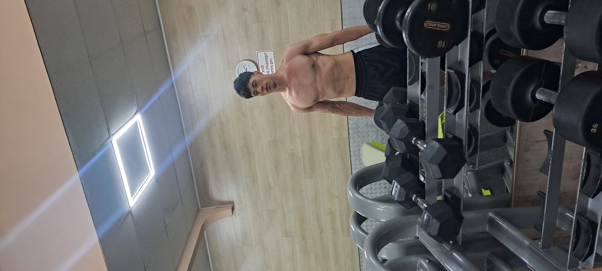 The image captures a muscular, lean man posing shirtless in a gym located in Burgas, Bulgaria. He stands with a neutral expression, looking towards the camera, displaying prominent vascularity in his arms. He is wearing black shorts with white side stripes.
To the man's right, a multi-tiered metal rack holds numerous black dumbbells, with some weights visible, such as "10" and "36", and the brand name "STAR TRAC". The gym floor features light wooden laminate transitioning to a darker, textured rubber mat in the area of the dumbbell rack.
On the wall behind the man, a round clock is partially visible, showing the time. Adjacent to the clock is a red-bordered sign with white text that appears mirrored and upside down; it seems to be a gym instruction, possibly stating "Сухи дрехи и обувки, моля!" (Dry clothes and shoes, please!). The ceiling consists of light grey acoustic tiles, with a bright square LED light fixture illuminating the space and casting distinct light rays. The overall scene suggests an indoor gym environment with clear, artificial lighting.