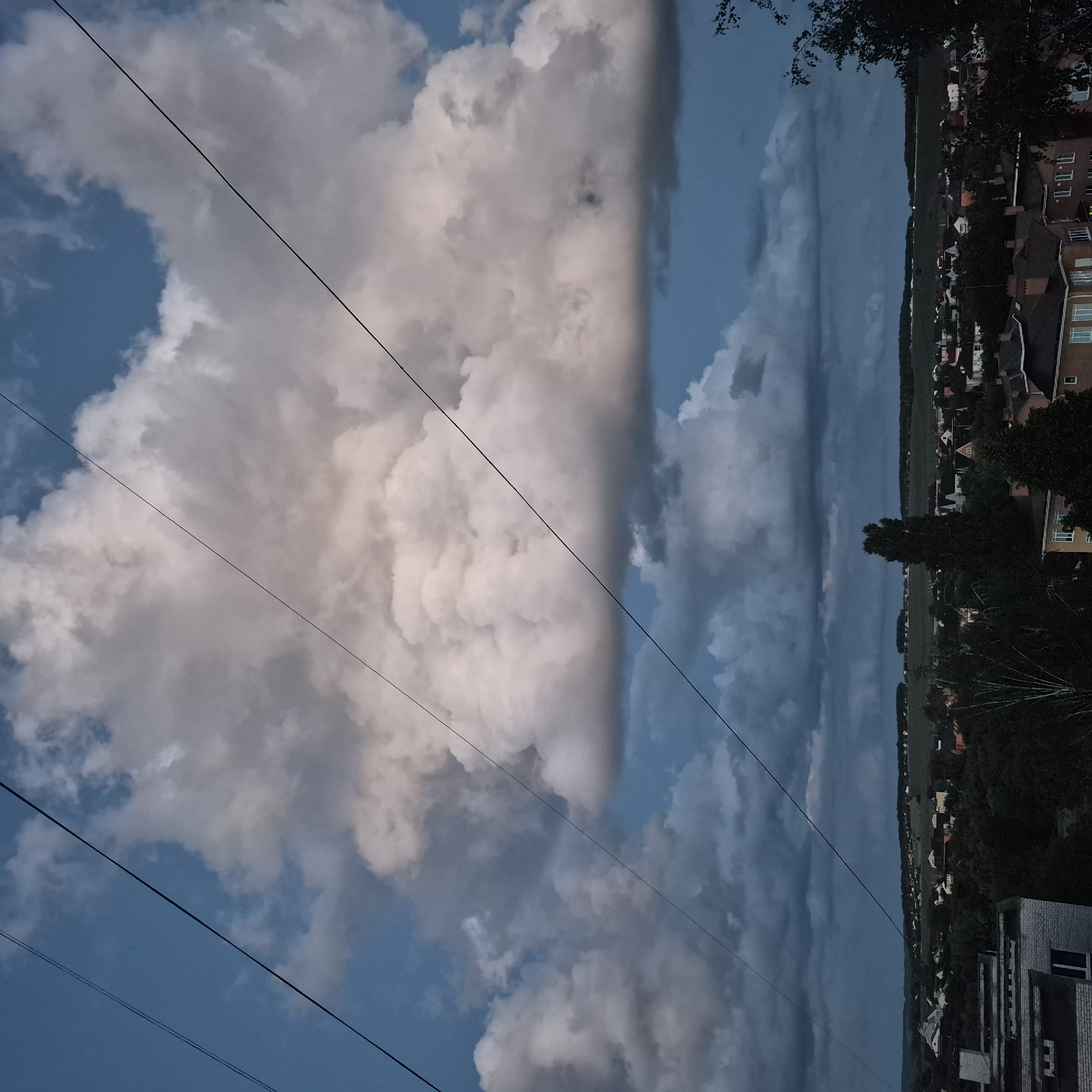 The image depicts a cloudy sky over a residential area, likely at dusk, with power lines stretching across the frame. The sky is a mix of deep blues and grays, with large, white cumulus clouds dominating the upper portion. Below the clouds, a town or village is visible, characterized by rooftops and trees. The scene appears to be taken from an elevated position, looking down upon the landscape. The lighting suggests it is either early morning or late evening. The presence of power lines indicates an urban or semi-urban setting.