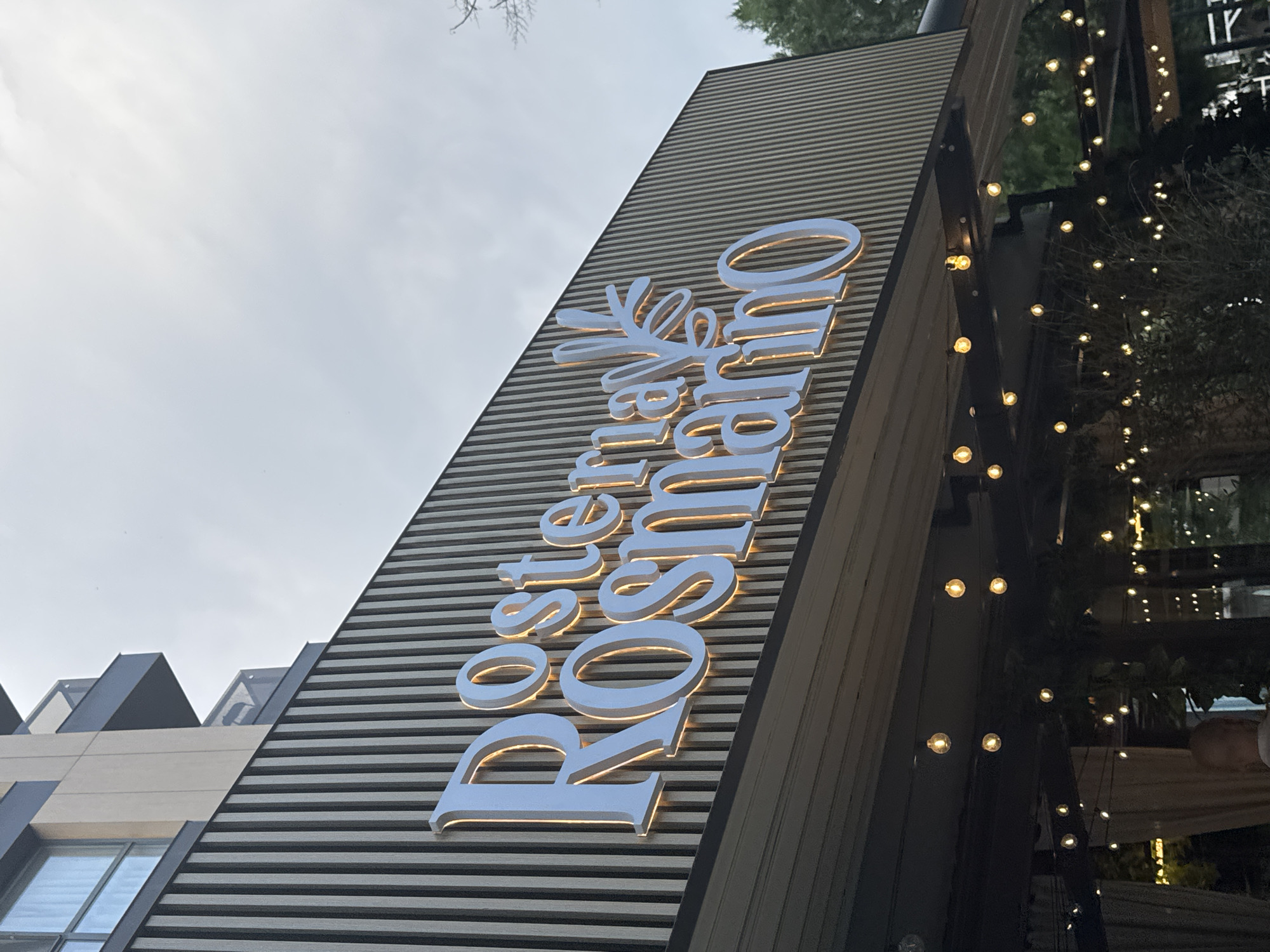 The image shows the illuminated signage of "Rostermay Rosmarino" on a building facade. The sign features large, white, 3D letters with a warm, orange glow emanating from behind them. The building has a modern design with vertical wooden slats on the exterior. Below the sign, an outdoor seating area is visible, decorated with strings of small, incandescent light bulbs that are also illuminated. The sky above is overcast. The establishment is located on the pedestrian street "Knyaz Alexander I" in Plovdiv, Bulgaria.