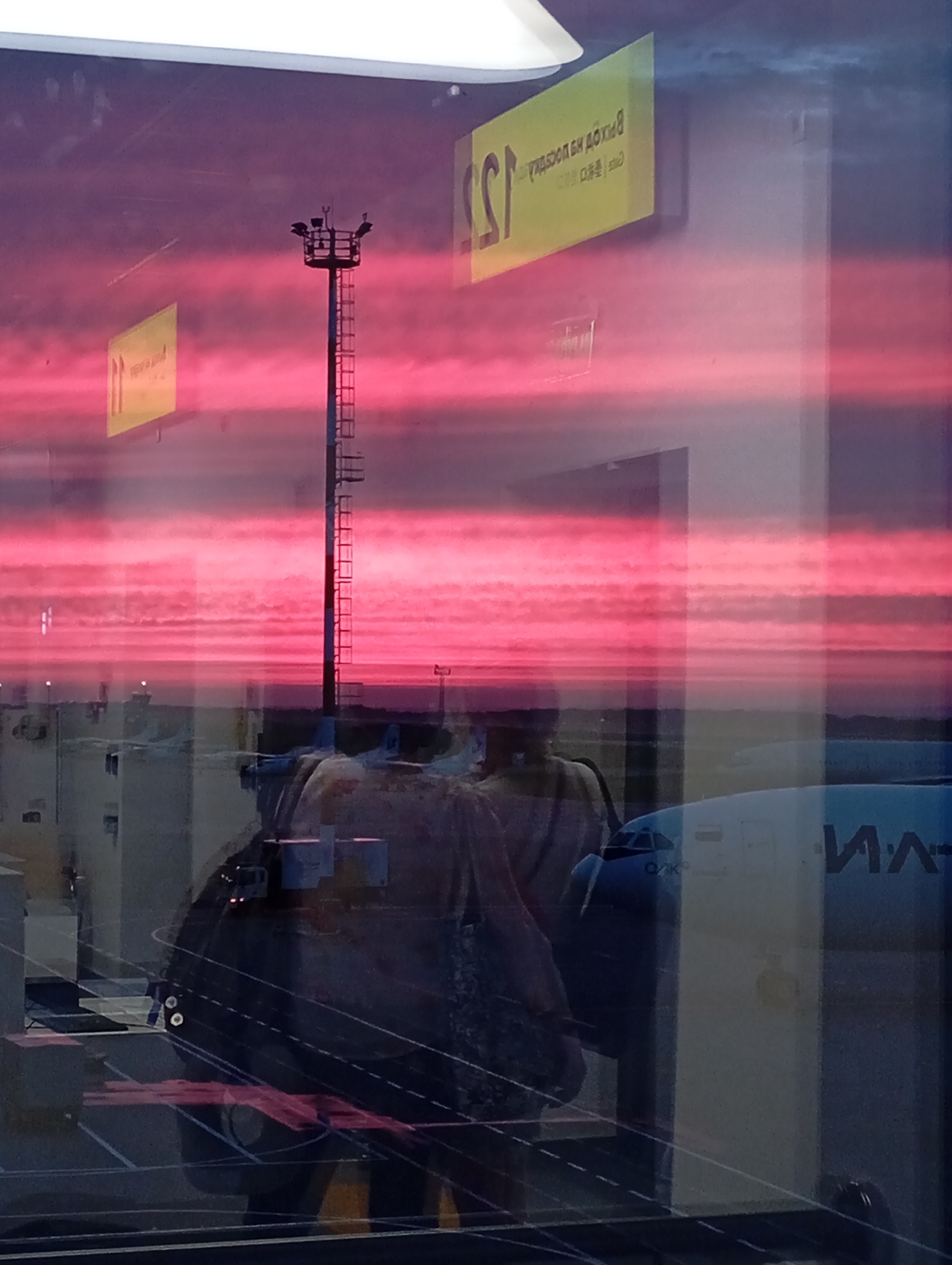 The image shows a reflection on a glass surface, likely a window at Khabarovsk Novy Airport in Khabarovsk, Russia. The reflection captures a vibrant pink and purple sunset sky, with horizontal bands of clouds. In the foreground, there are reflections of people, possibly passengers or airport staff, and parts of an airplane. A tall tower with lights, likely an airport floodlight, is also reflected. Yellow signage with Cyrillic text is visible in the upper portion of the reflection, appearing as reversed lettering. The overall scene suggests an atmospheric and somewhat surreal view of an airport during sunset.