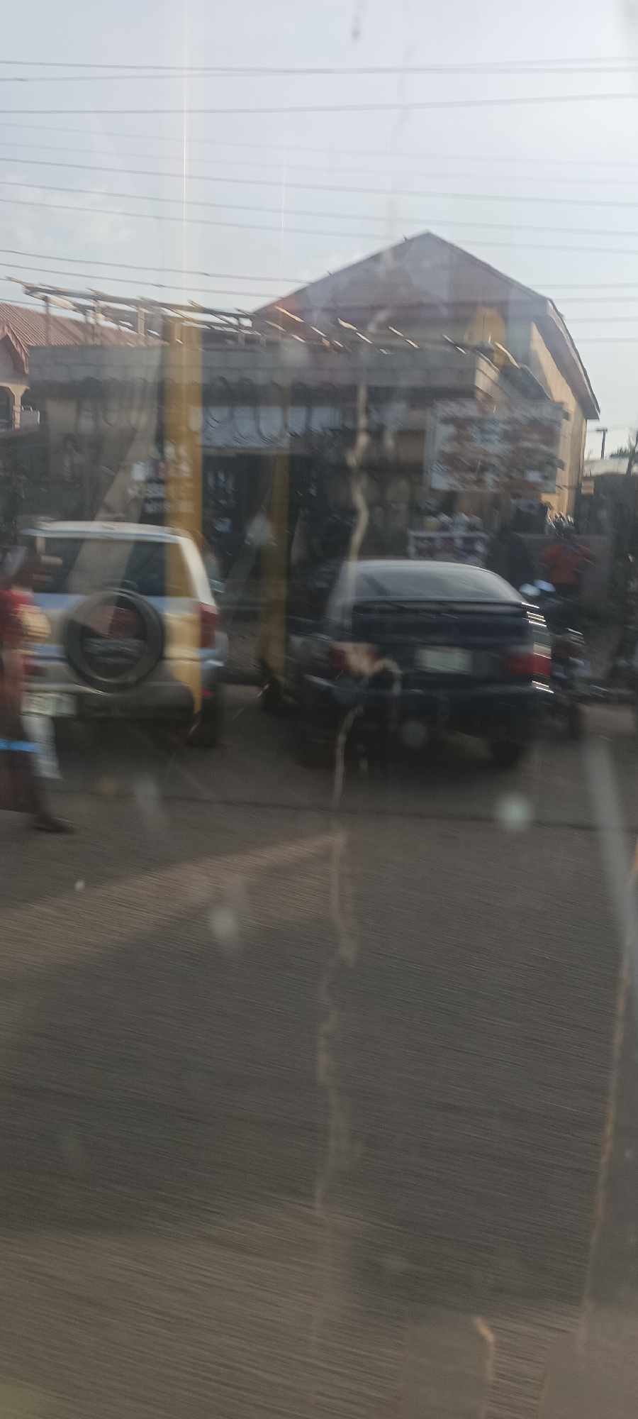 This media file depicts a daytime street scene in Gura Top, Nigeria, likely photographed from inside a vehicle, as evidenced by visible reflections and some blurring on the glass surface. The bright, clear sky indicates fair weather.
In the mid-ground, a row of buildings lines the street, appearing to be commercial establishments or residences. These structures feature modest architecture, with some showcasing corrugated metal roofing and one prominent building featuring a gabled roof. Power lines crisscross the sky above.
On the street, a light-colored SUV with a spare tire mounted on its rear is visible on the left side, with partially discernible text on its tire cover. A dark-colored sedan is parked to its right, and a motorcycle or scooter is partially visible behind the sedan. A person's lower body is visible on the far left, suggesting pedestrian movement.
The foreground shows a paved road surface with indistinct markings or cracks. The overall scene captures a typical, bustling street environment.