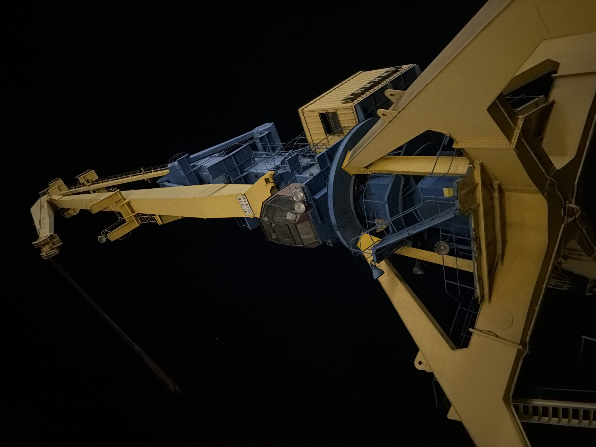 An upward-angle shot shows a large crane against a black night sky. The crane is predominantly yellow and blue, with a complex lattice-like structure visible. The crane's boom extends to the left. The operator's cabin is visible with windows. A cable descends from the boom into the darkness. The location is likely Burgas, Bulgaria, due to the presence of such cranes in port or shipyard areas. No people are visible.
