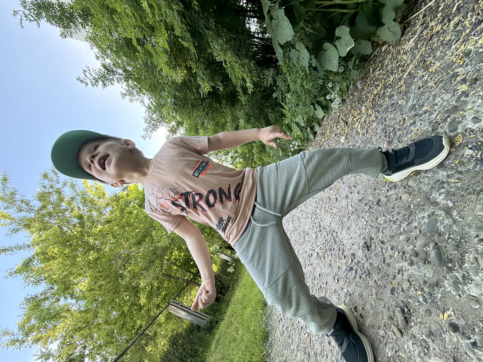 A child wearing a green baseball cap, a pink t-shirt with the word "STRONG" on it, and grey sweatpants is standing on a gravel path. The child is mid-stride, with one leg extended outwards, giving the impression of movement. Trees and foliage are visible in the background, suggesting an outdoor setting. The gravel path extends into the distance, and part of what appears to be a metal object is visible on the left side of the frame. The angle is low, looking upwards at the child. The location is Chaikovsky, Russia.