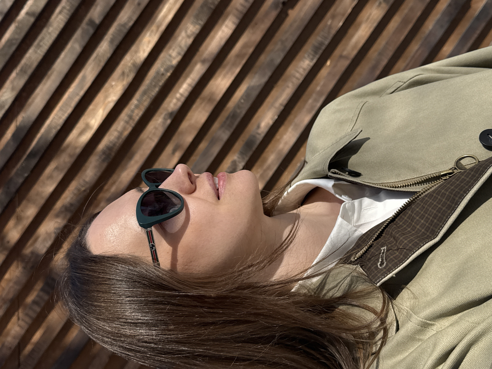 A woman is reclining outdoors, wearing sunglasses and a light-colored jacket over a white shirt. She appears to be on a wooden structure, possibly a bench or platform, with a slatted wooden background. The image does not contain sufficient information to ascertain the location in Chaikovsky, Russia.