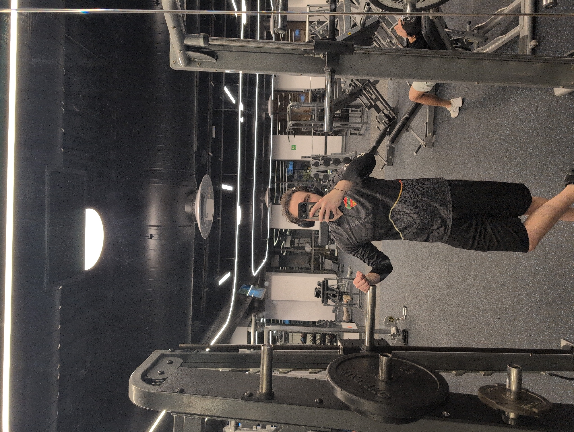 A male is taking a mirror selfie in a dimly lit gym. He is wearing headphones, a black long-sleeve shirt with a gray, yellow, and red pattern on the chest, and black shorts. He is standing in front of a weight machine with large circular weights attached to it. In the background, another person is visible lying on a weight machine. The gym has dark ceilings with modern lighting fixtures and a black rubber floor. The mirror reflects the gym equipment and lights. There is no indication that the image was taken in Mexico City, Mexico.