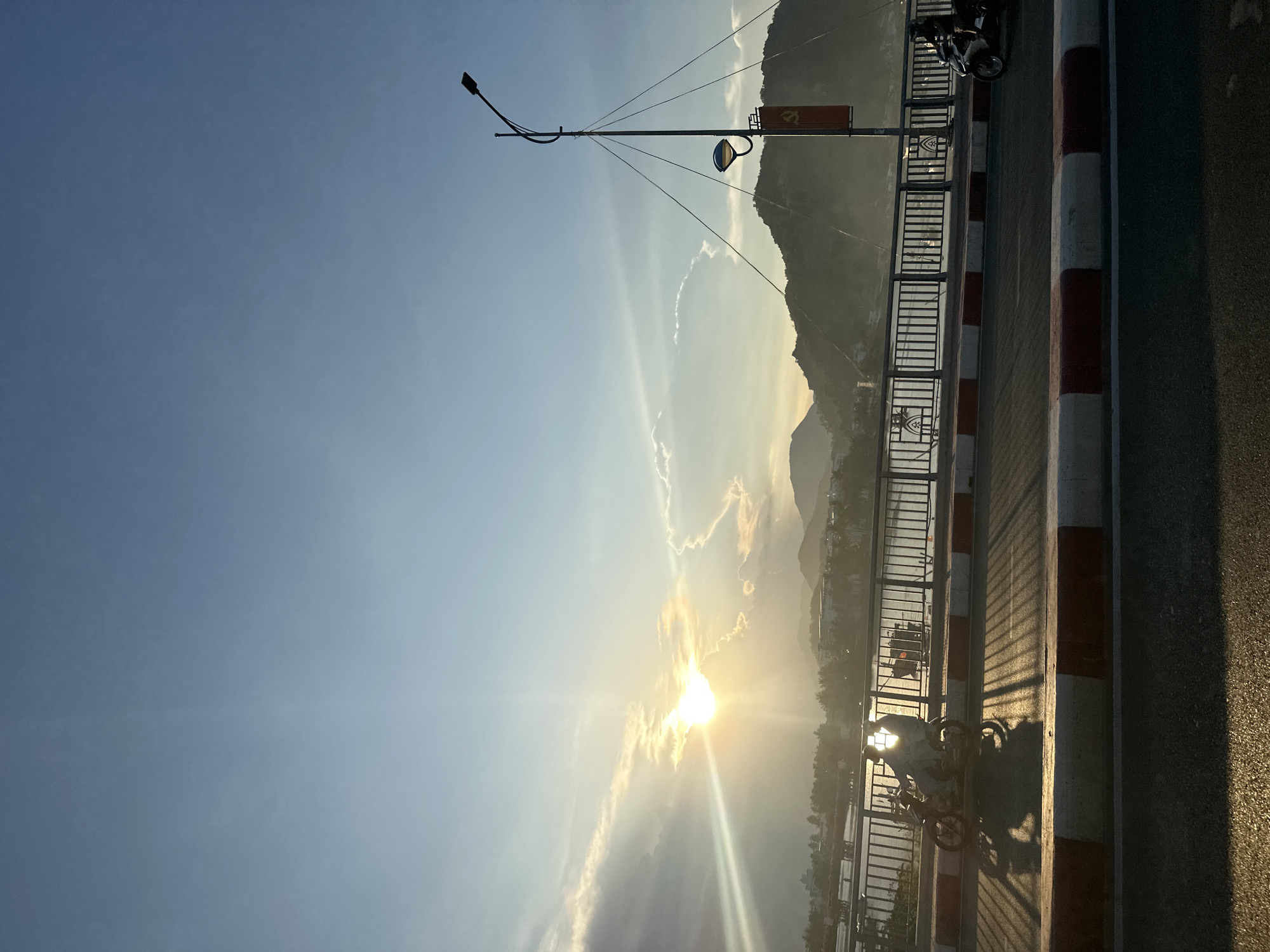 A bright, sunny day on a bridge with the sun low on the horizon, likely in the city of Nha Trang, Vietnam. The sky is a clear blue with some wispy clouds near the sun. The sun's rays are prominent, casting a strong glare and silhouetting the landscape. The bridge has a striped red and white barrier. A motorcyclist is on the bridge. A streetlight and other infrastructure are visible, along with some mountains in the background. The photo is oriented vertically.