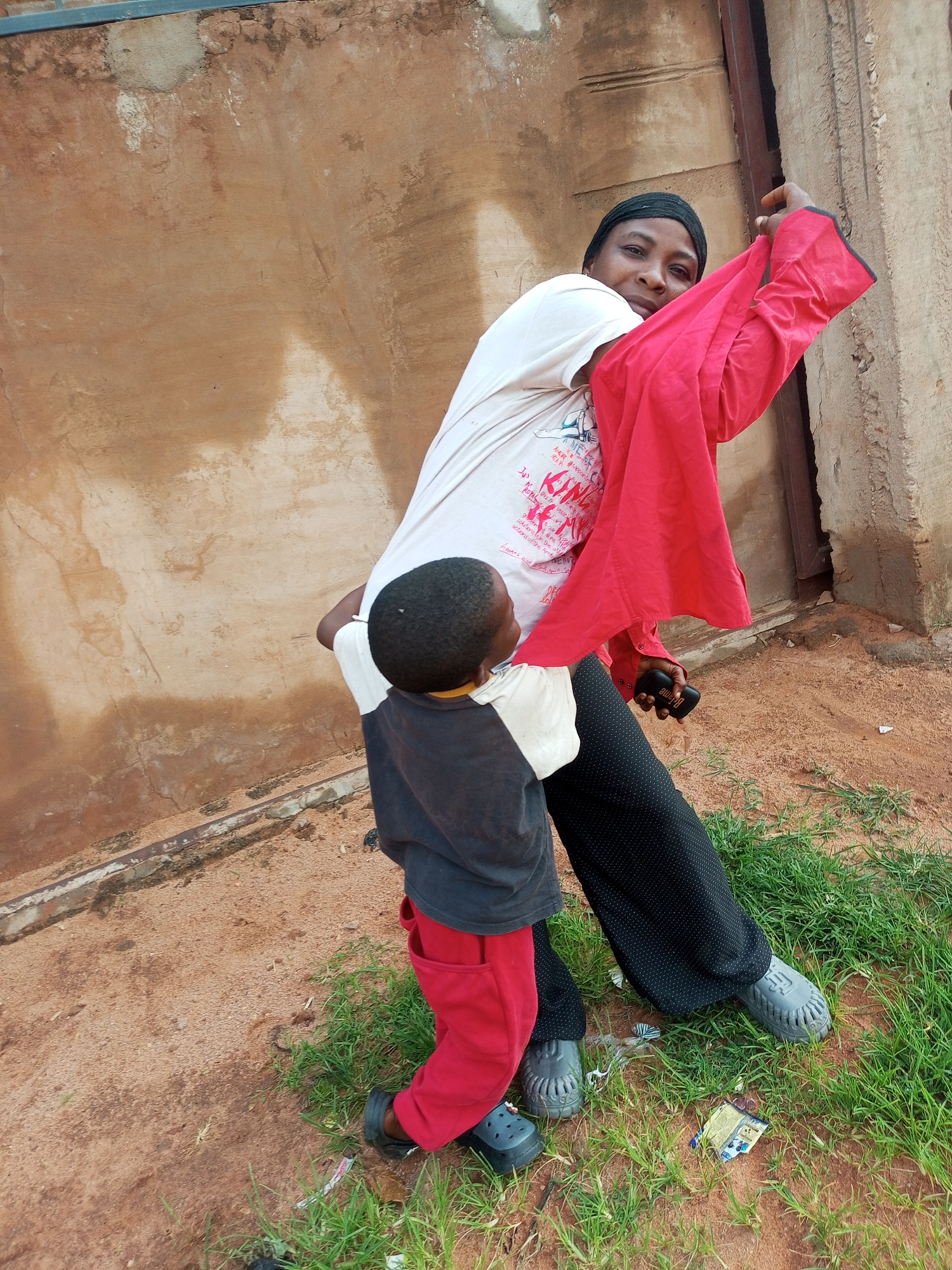 In Tempure, Nigeria, a woman wearing a black head covering, a white t-shirt, and black patterned pants stands outdoors. She is holding a bright red garment over her shoulder and appears to be interacting with a small child. The child, wearing a grey and black shirt with red pants, is positioned in front of the woman and looking up at her. They are standing on a ground covered with sand and patches of green grass. Behind them is a wall with a rough, textured surface in shades of brown and beige, with some lighter patches and darker staining. A dark vertical element, possibly a door or frame, is visible on the right side of the image. The lighting suggests it is daytime. The woman is holding a black rectangular object in her right hand.