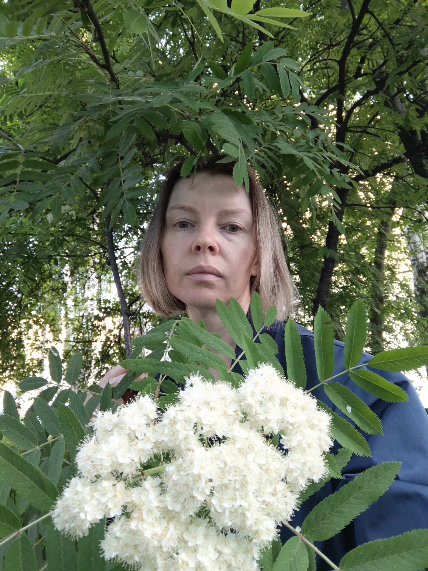 A fair-skinned woman with short, light brown hair is centered in the frame, holding a large cluster of white flowers and leafy green foliage in front of her chest. She has a neutral expression and is wearing a dark blue garment. The background is filled with dense green leaves and tree branches, suggesting a forest or park setting. Sunlight filters through the leaves, creating a dappled effect. The overall scene appears to be a natural outdoor environment. This photo was taken in the city of Chaikovsky, Russia.