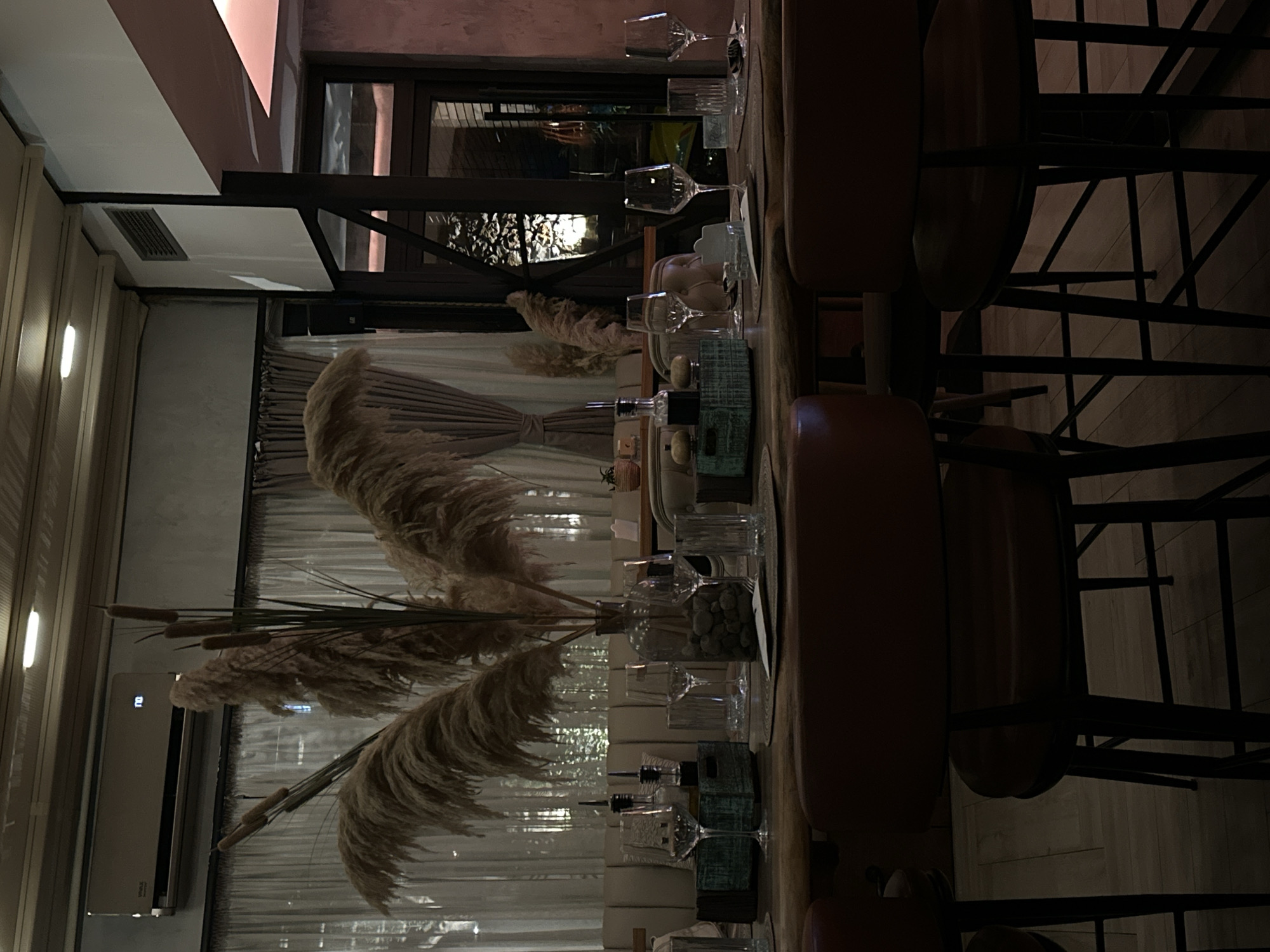 The image depicts the interior of a dimly lit restaurant in Šušanj, Montenegro. The foreground is dominated by a wooden table set for dining with several wine glasses, cutlery, and placemats. A large vase filled with dried pampas grass and pebbles serves as a centerpiece. In the background, a beige upholstered banquette seating area is visible, separated from the dining area by curtains. An air conditioning unit is mounted on the wall above the banquette. Through a large window or glass door in the background, another part of the restaurant with a warm, ambient lighting is visible, suggesting a sophisticated dining atmosphere. The overall scene is characterized by warm tones and natural textures, creating an inviting ambiance. There are no people visible in the image.